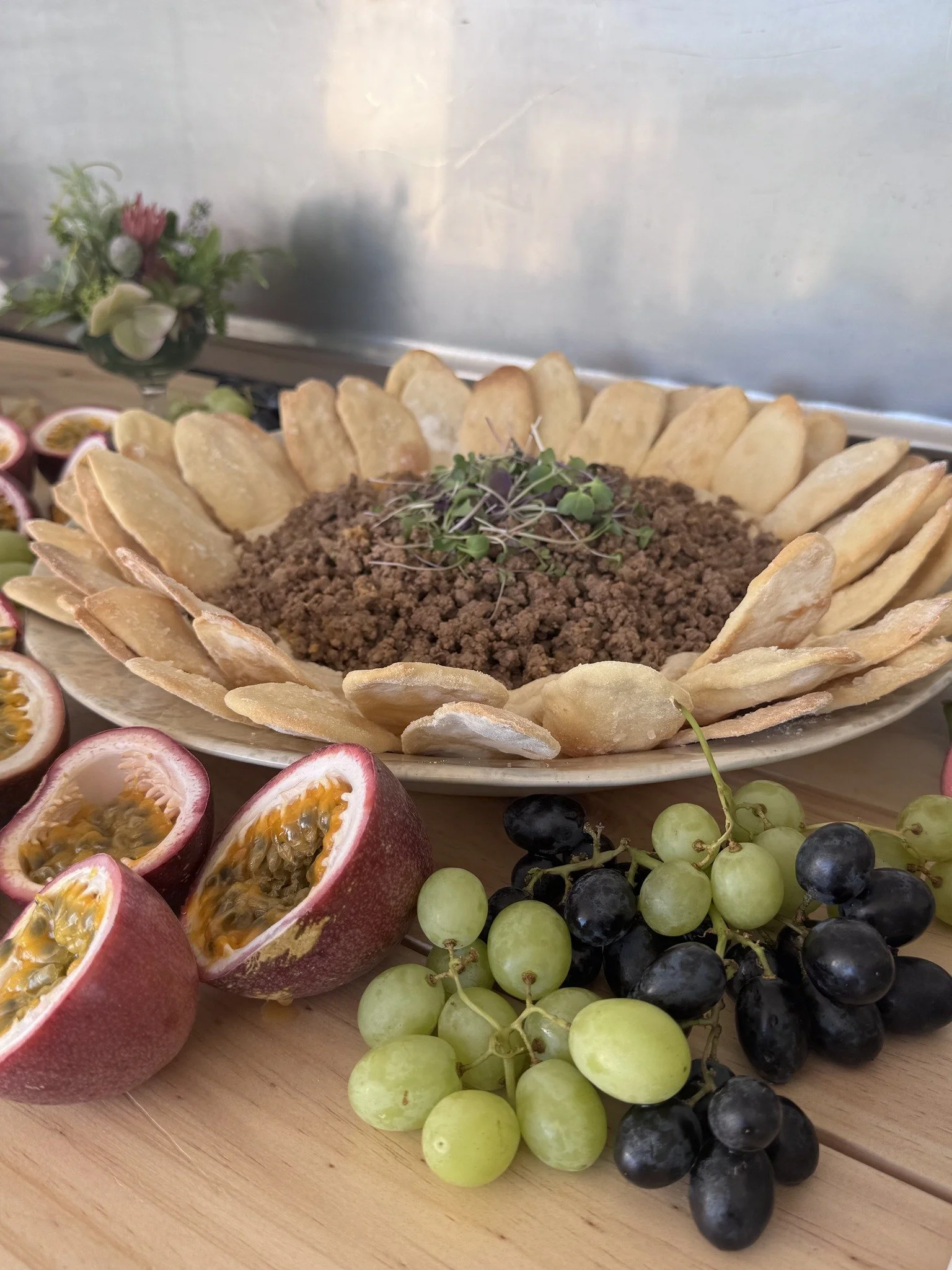 Plate of ground beef topped with microgreens, surrounded by potato chips, with halved passion fruits and green and black grapes in front.