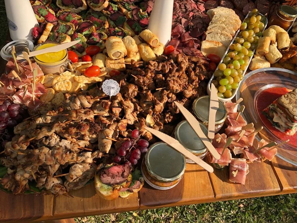 Variety of appetizers including grilled skewers, cheese, crackers, cherry tomatoes, green grapes, a layered sandwich, and spreads or dips, arranged on a wooden table.