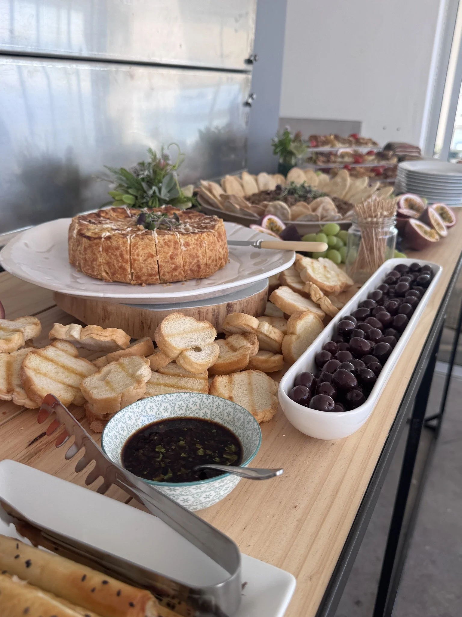A buffet table with various cheeses, bread, crackers, grapes, olives, and dipping sauces, including a round cheese wheel garnished with herbs.