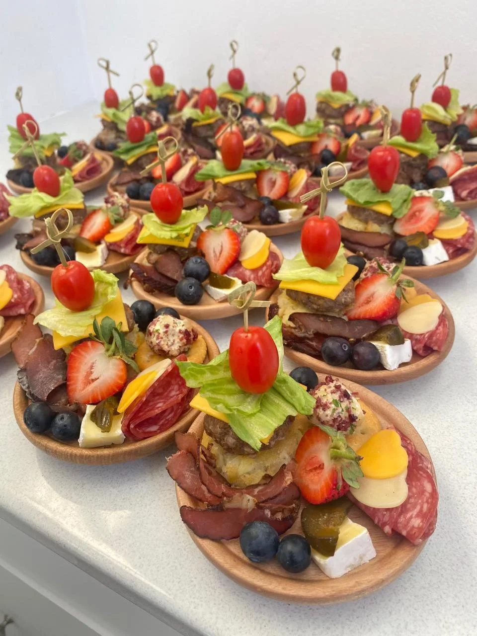 Assorted appetizer bites on small wooden spoons, featuring cherry tomatoes, lettuce, cheese, sliced meats, strawberries, blueberries, pickles, and crackers, garnished with small wooden skewers.