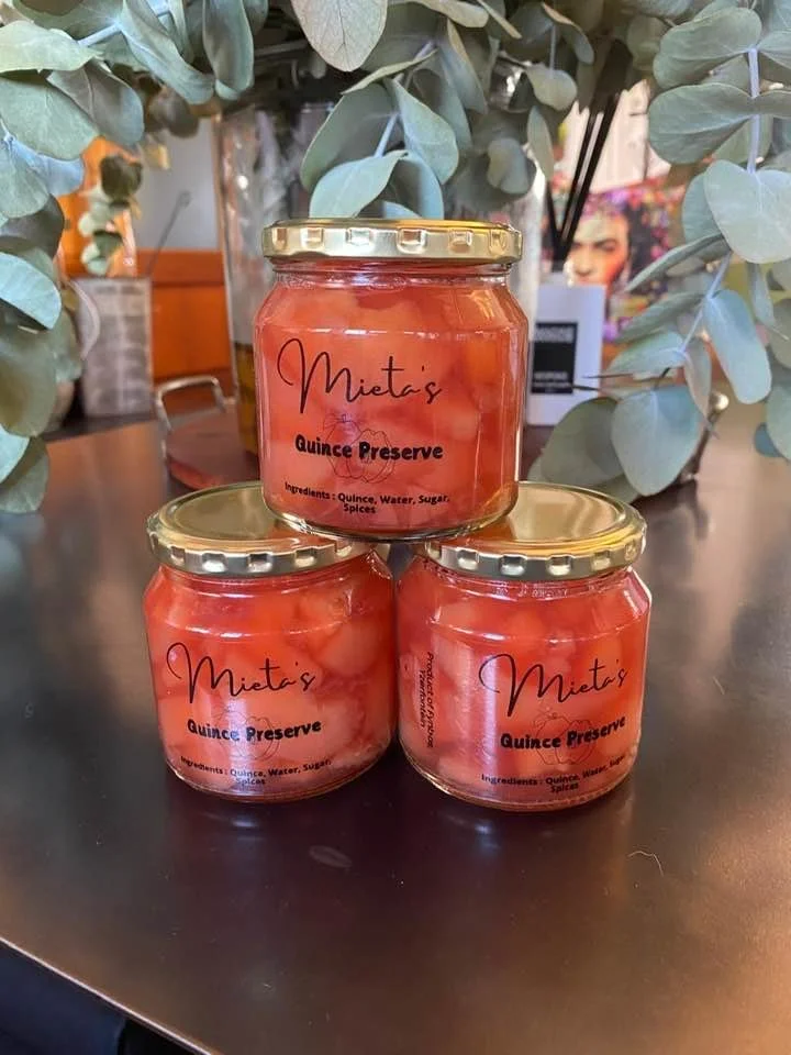 Three jars of quince preserve placed on a dark surface with eucalyptus leaves in the background. The jars have gold lids and labels that read 'Mieta's Quince Preserve,' and list ingredients as quince, water, sugar, and spices.