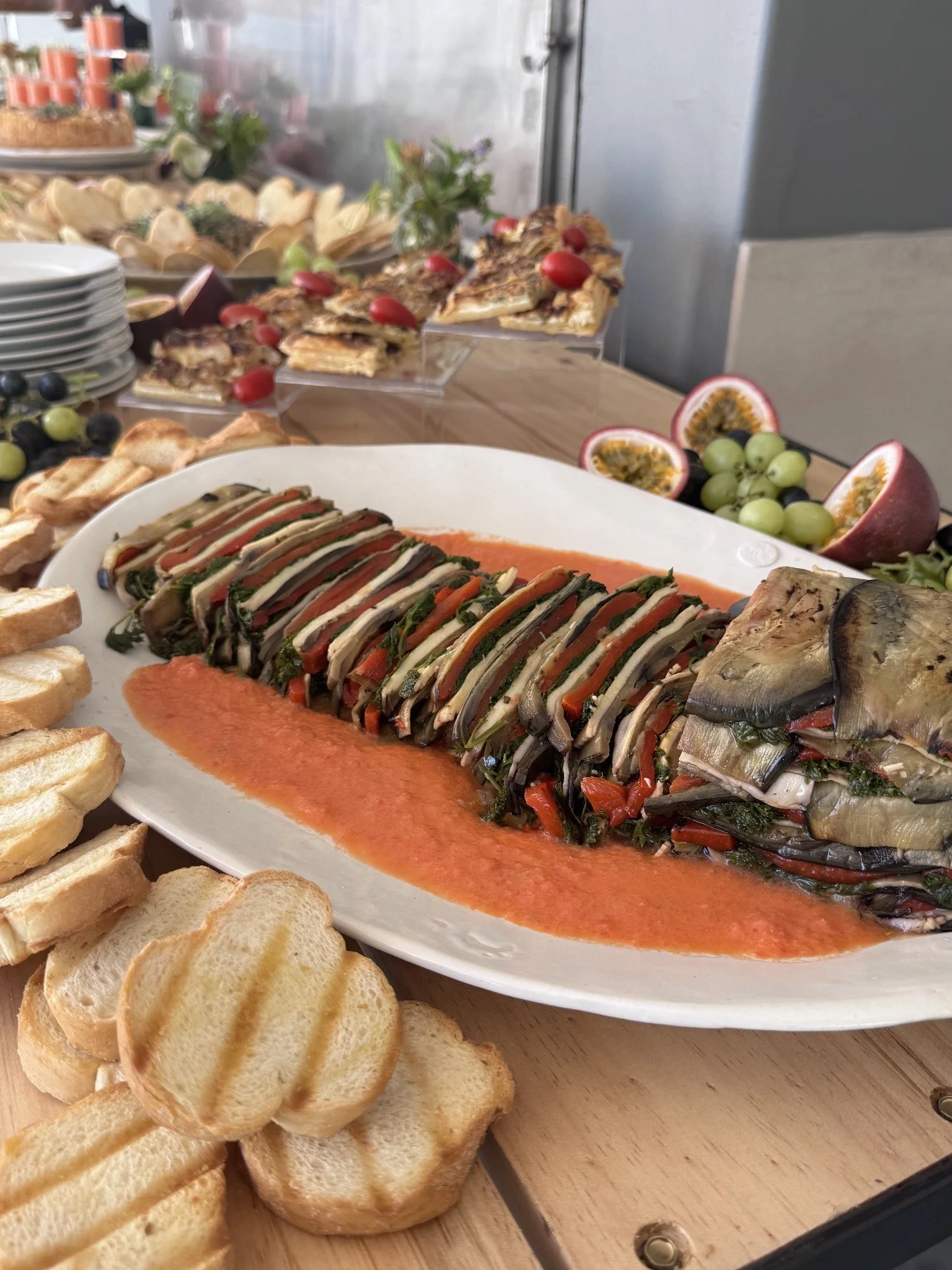 A seafood dish with sliced eggplant, peppers, and greens in a red sauce, garnished with smaller vegetables, served with toasted bread slices. In the background, there are additional dishes including a pizza topped with cherry tomatoes, and fruit such