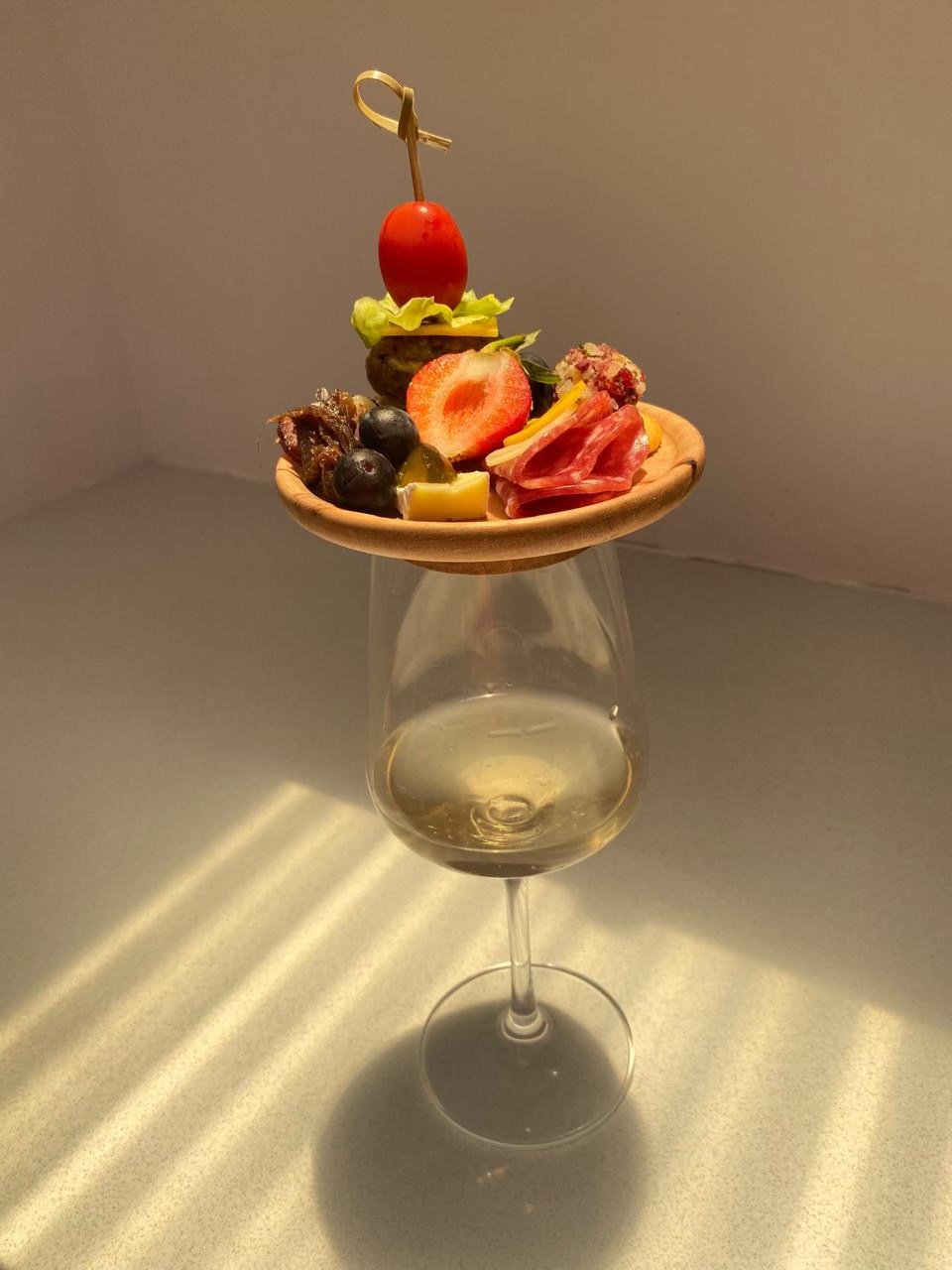An oil glass with water, placed on a surface in front of a plain wall. On top of the glass, there is a small wooden plate with various fruits, vegetables, and meats arranged on it.