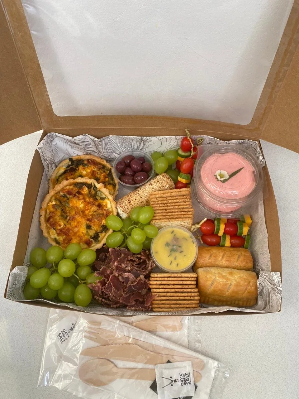A takeout box with two mini pizzas, green grapes, red grapes, cherry tomatoes on skewers, a container of pink dessert, a container of yellow sauce, two breaded rolls, crackers, beef jerky, and a few other snacks.