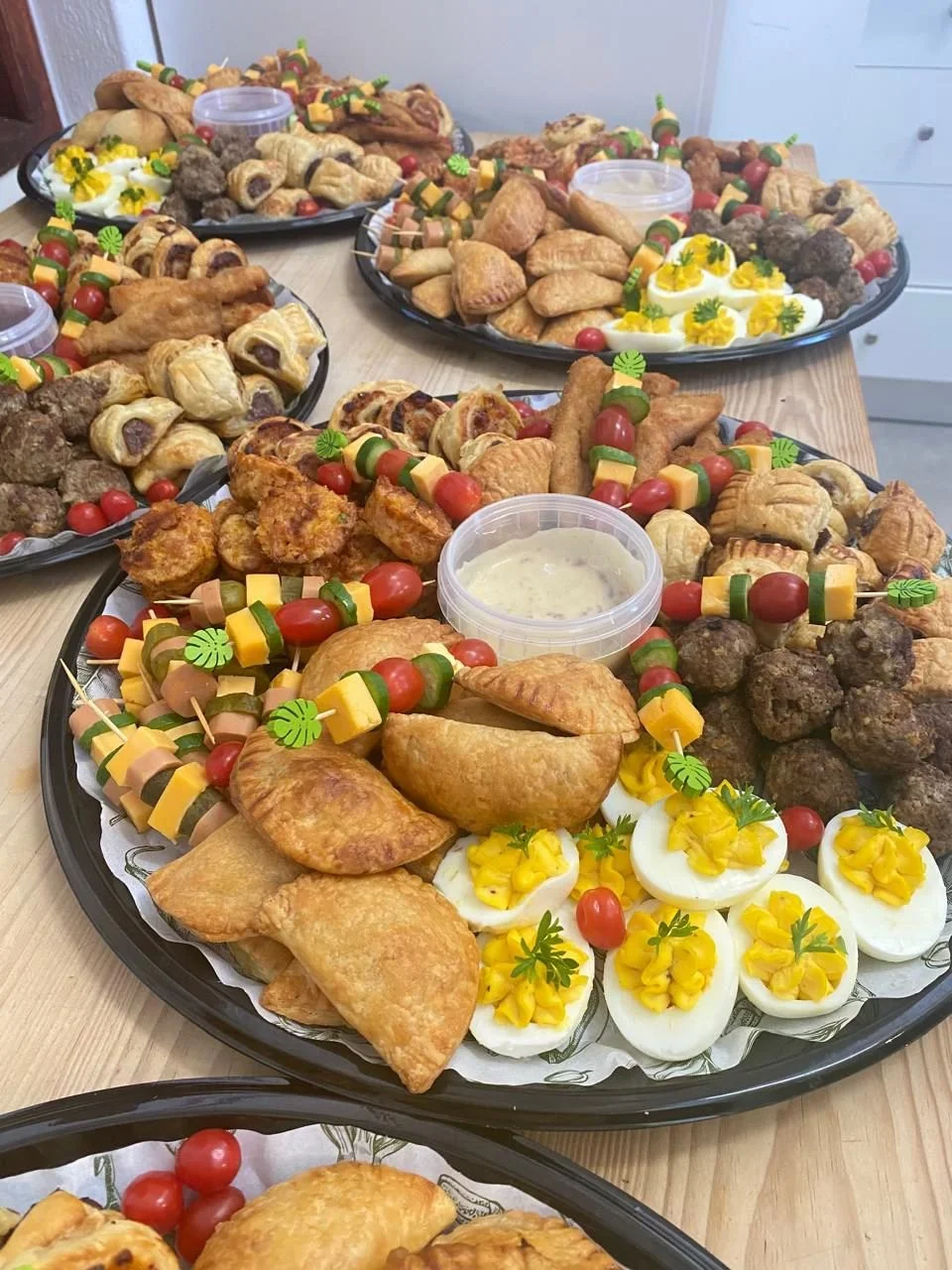 Large dinner platters filled with various appetizers including deviled eggs topped with yellow peppers, cherry tomatoes on skewers with cheese and vegetables, fried chicken drumsticks and other fried snacks, meatballs, sausage rolls, and small sandwi