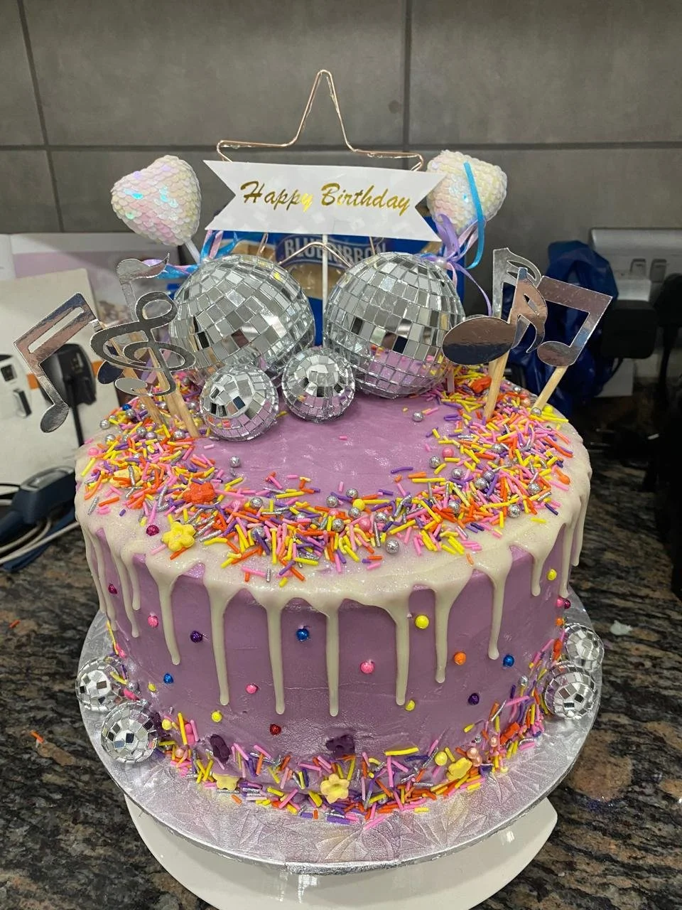 A purple birthday cake with white dripping icing, decorated with colorful sprinkles, silver ball and disco ball decorations, and musical note toppers, with a "Happy Birthday" sign on top.