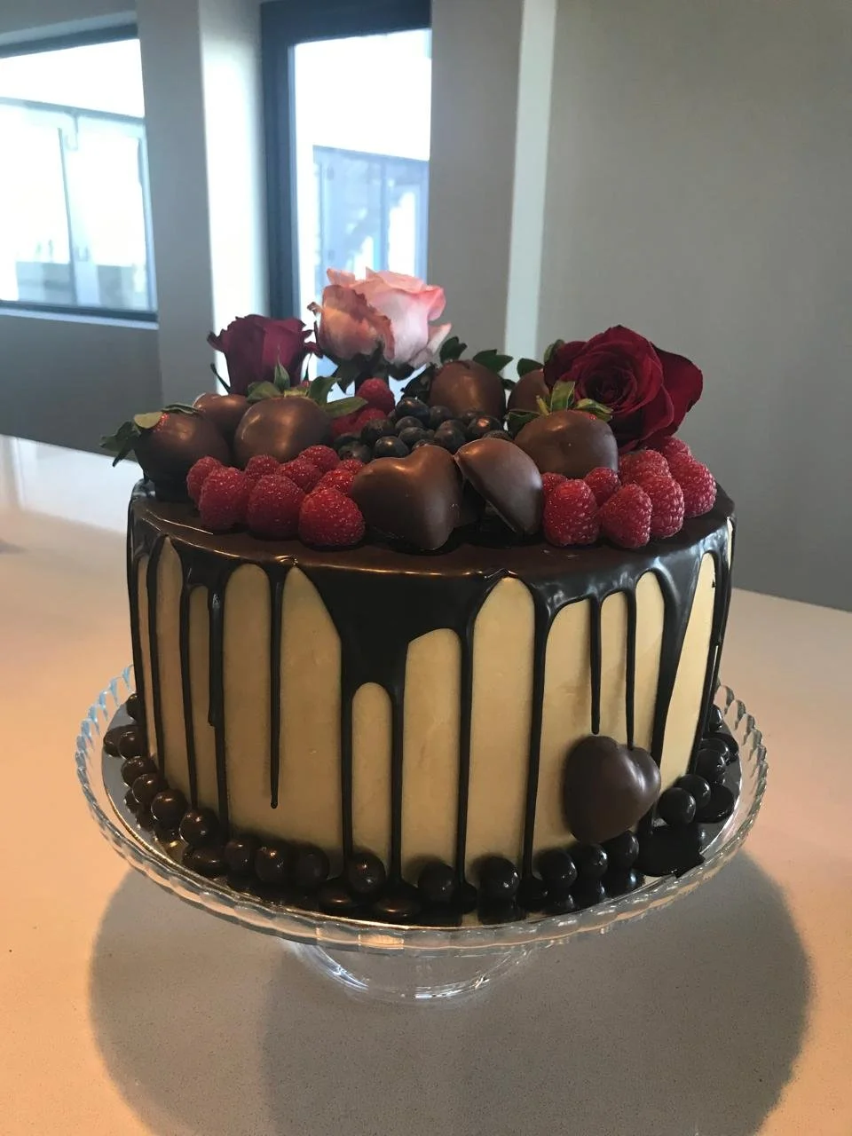 A decorated cake with white frosting, topped with red and pink roses, raspberries, chocolate-covered strawberries, and blueberries, with chocolate drizzles on top and a border of blueberries at the base.