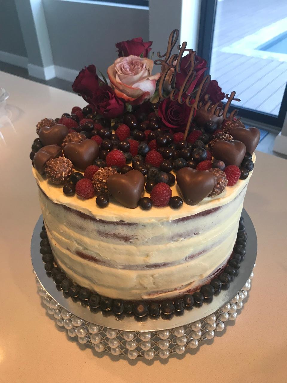 A decorated birthday cake with a semi-transparent frosting, topped with chocolate heart-shaped candies, raspberries, blueberries, and a flower arrangement with a 'Happy Birthday' topper. The cake sits on a pearl-edged cake board.