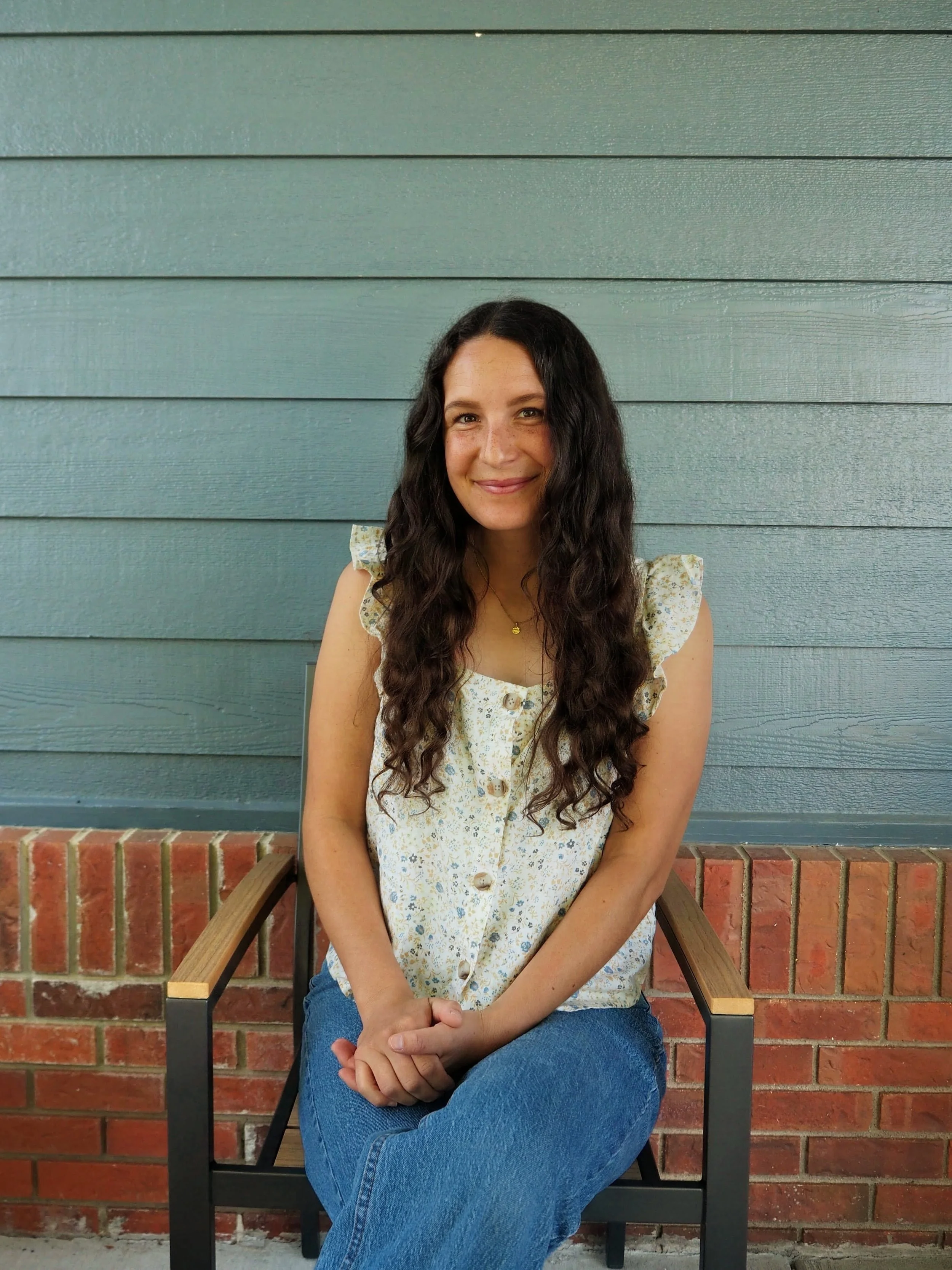 A woman with long dark wavy hair, wearing a cream-colored floral dress with buttons, sitting on a black chair with wooden armrests in front of a teal wooden wall and a red brick ledge.