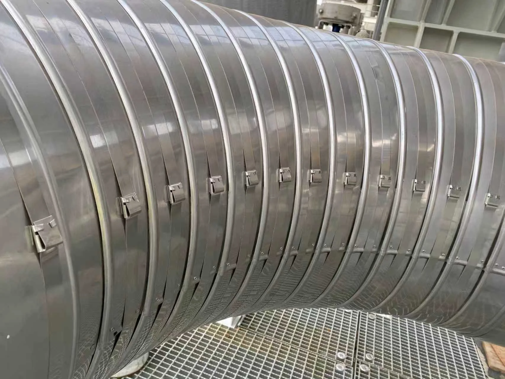 Close-up of a large, metallic, industrial pipe with multiple clamps securing the sections, situated above a grated platform in a factory or industrial setting.