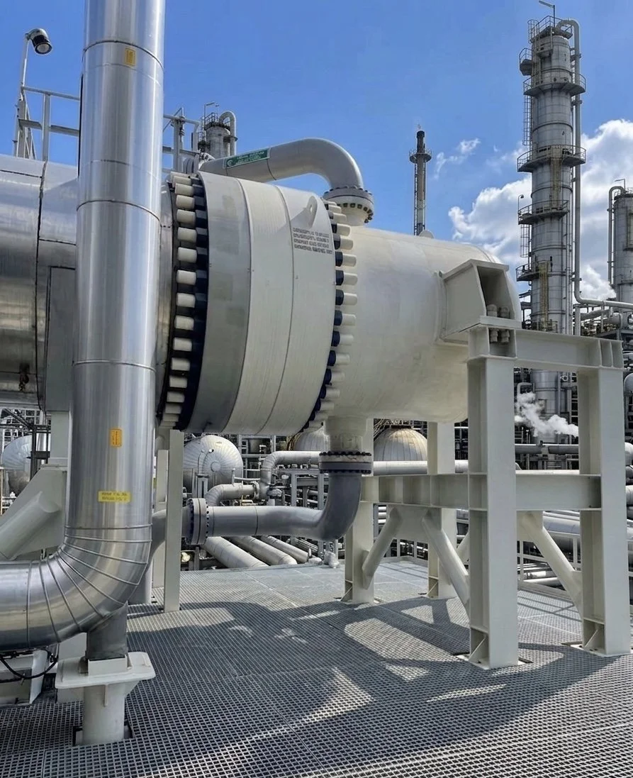 Large industrial equipment with pipes, valves, and tall towers at an industrial plant under a partly cloudy sky.