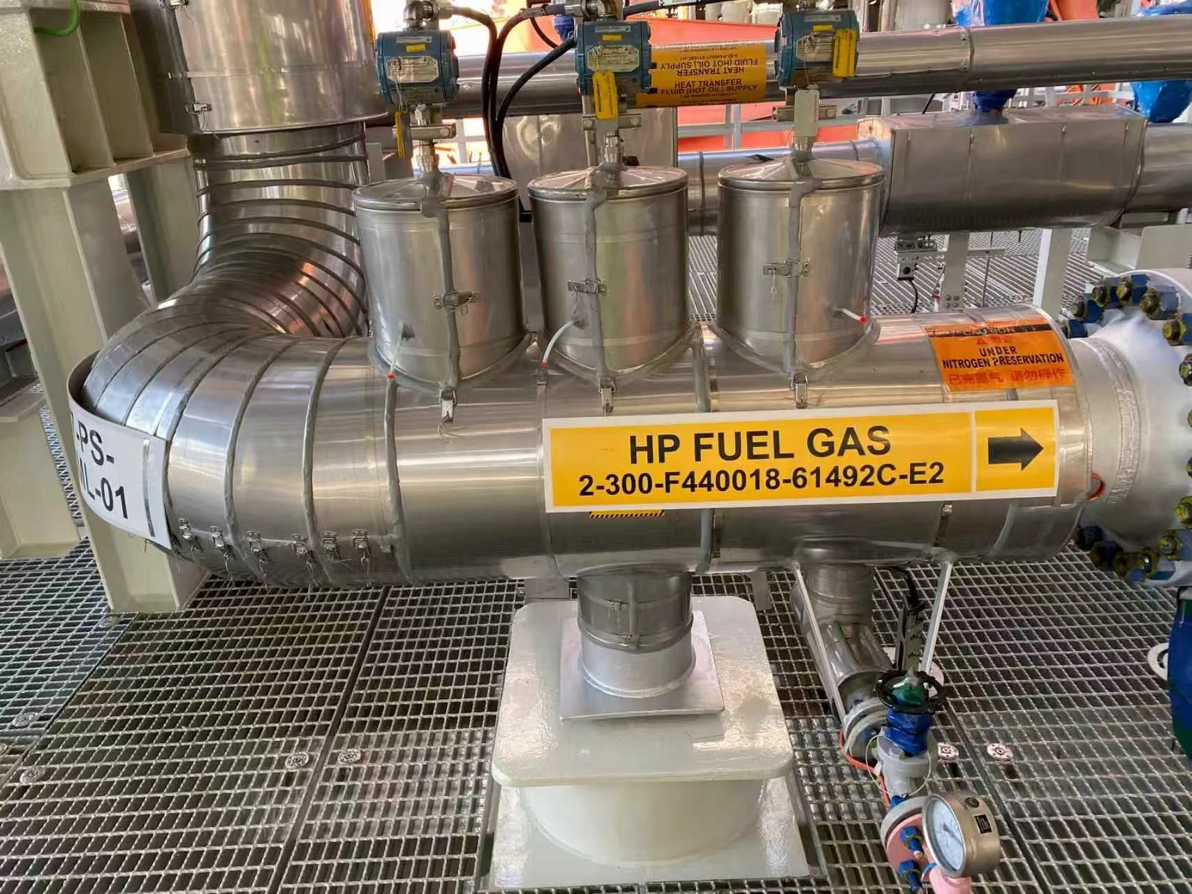 Industrial pipeline system with a large horizontal metal pipe labeled "HP FUEL GAS" and a yellow label, multiple smaller vertical cylinders attached, gauges, and warning labels, situated on a grated metal floor.