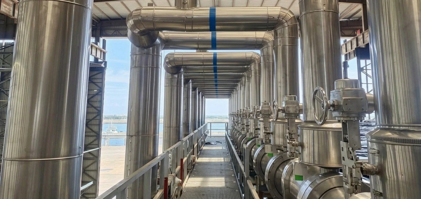 Industrial piping with large stainless steel pipes and valves on a walkway inside a facility, overlooking a water body outside.