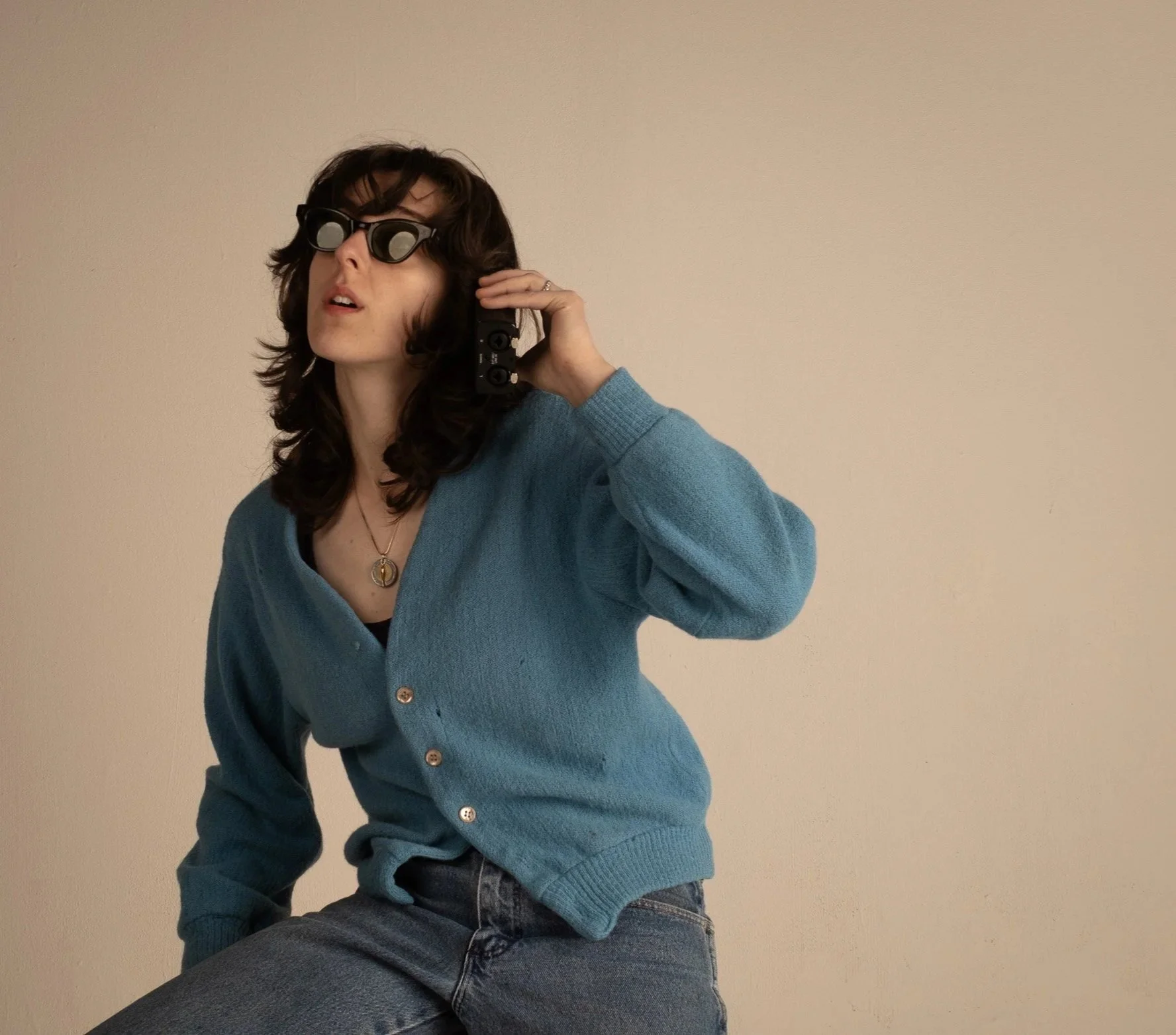 A young woman with dark, wavy hair wearing sunglasses, a blue cardigan, and jeans, taking a mirror selfie with a camera phone, against a plain beige wall.