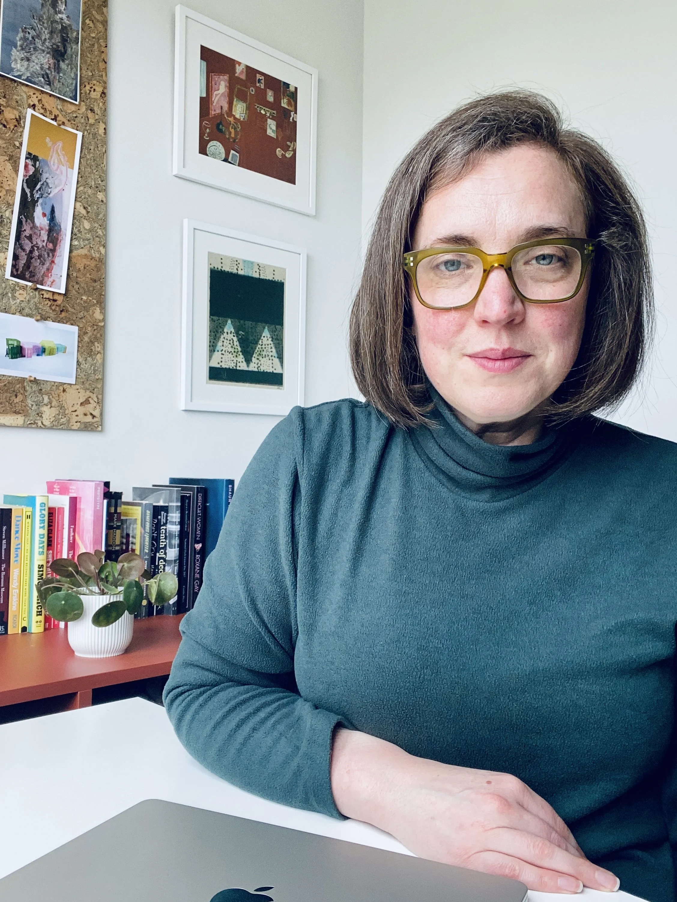 A woman with shoulder-length brown hair, light skin, and green glasses sitting at a desk, wearing a teal long-sleeve shirt, in a room with framed artwork on the wall and a bookshelf with colorful books.