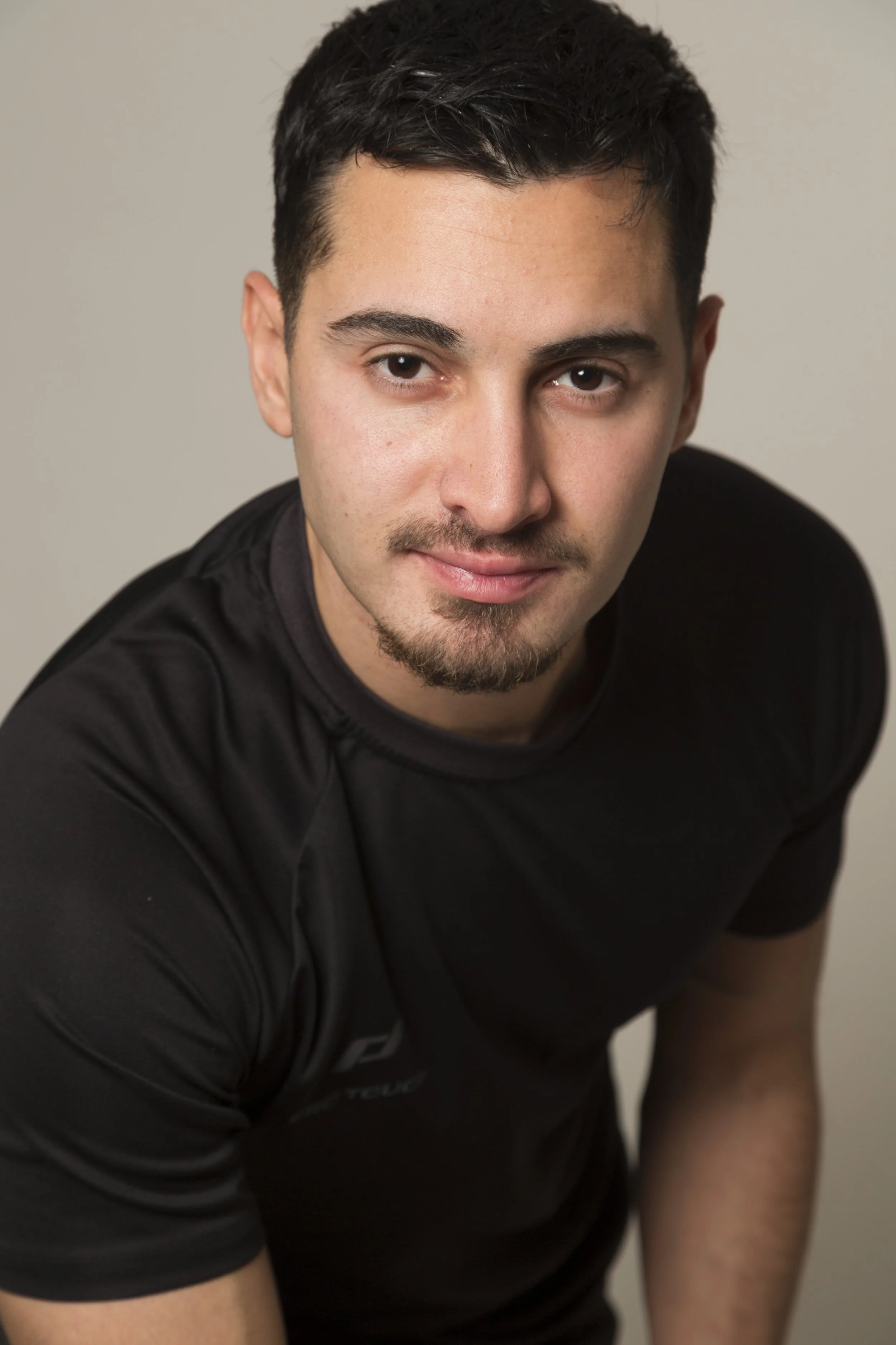 Portrait d'un jeune homme aux cheveux noirs, portant un t-shirt noir, regardant vers l'objectif avec un sourire léger dans une lumière douce.