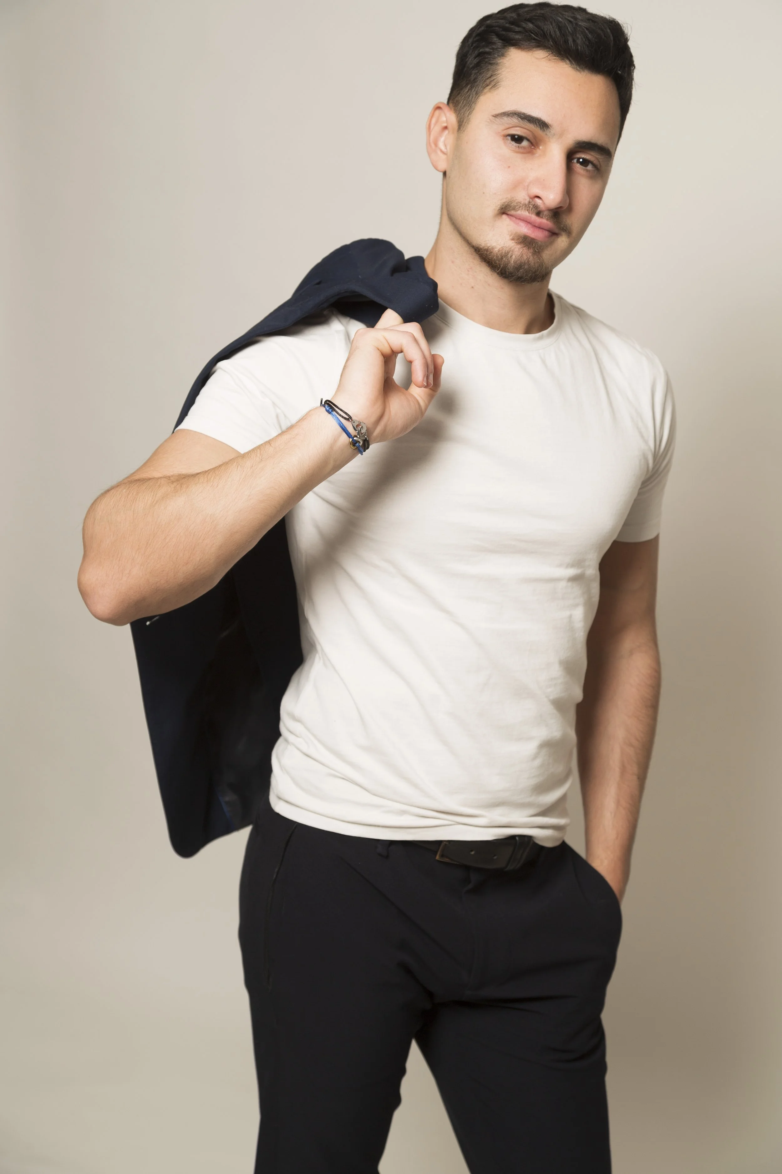 Jeune homme avec une barbe portant un t-shirt blanc, un pantalon noir et une veste noire sur l'épaule, posant de manière décontractée contre un mur beige.
