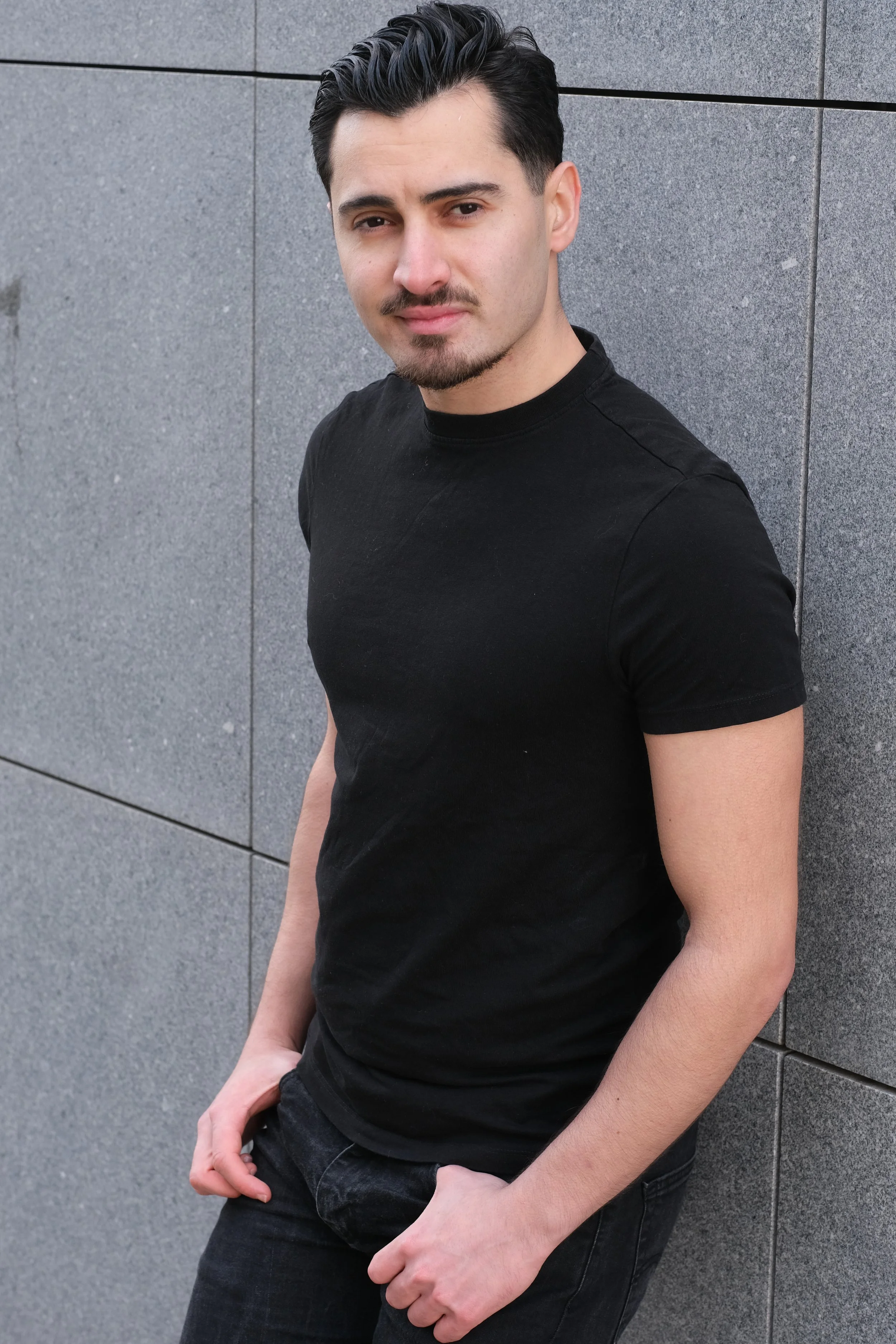 Un jeune homme avec des cheveux noirs, une moustache et une barbe, portant un t-shirt noir, posé contre un mur en pierre grise.