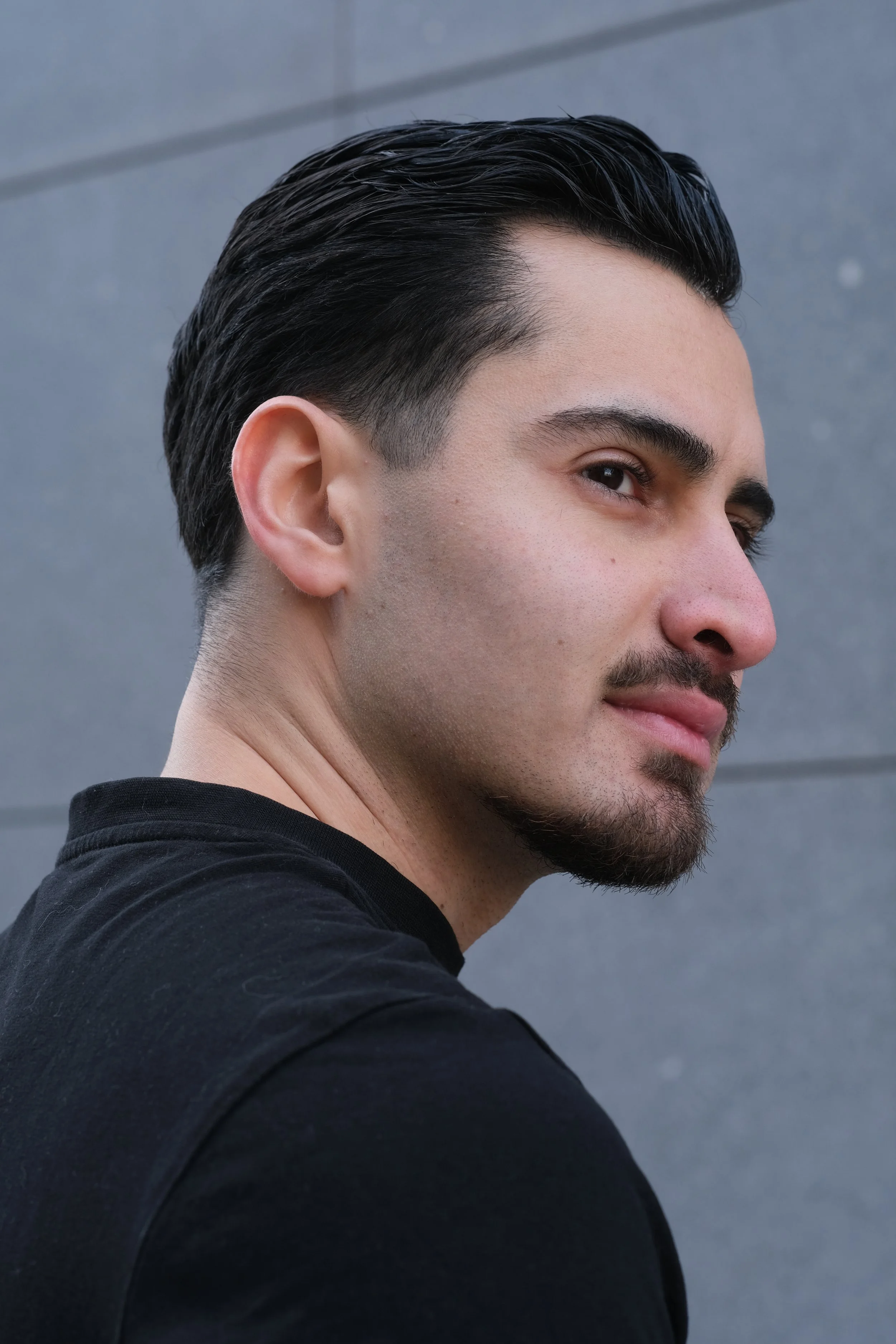 Close-up d'un homme avec les cheveux noirs coiffés en arrière, portant un t-shirt noir, contre un mur gris.