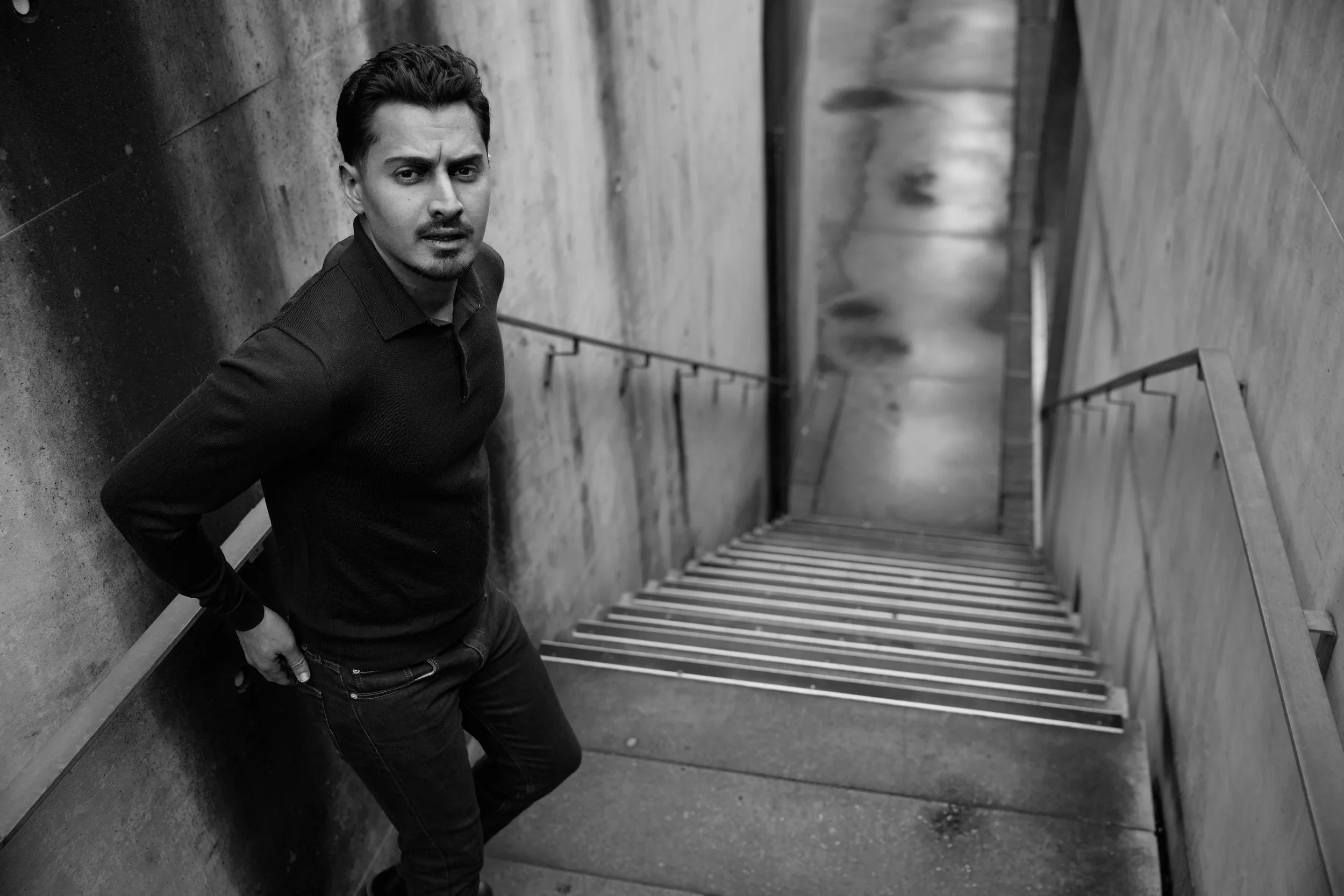 Homme debout dans une station de métro moderne, face caméra, avec murs en béton et escaliers mécaniques en arrière-plan.