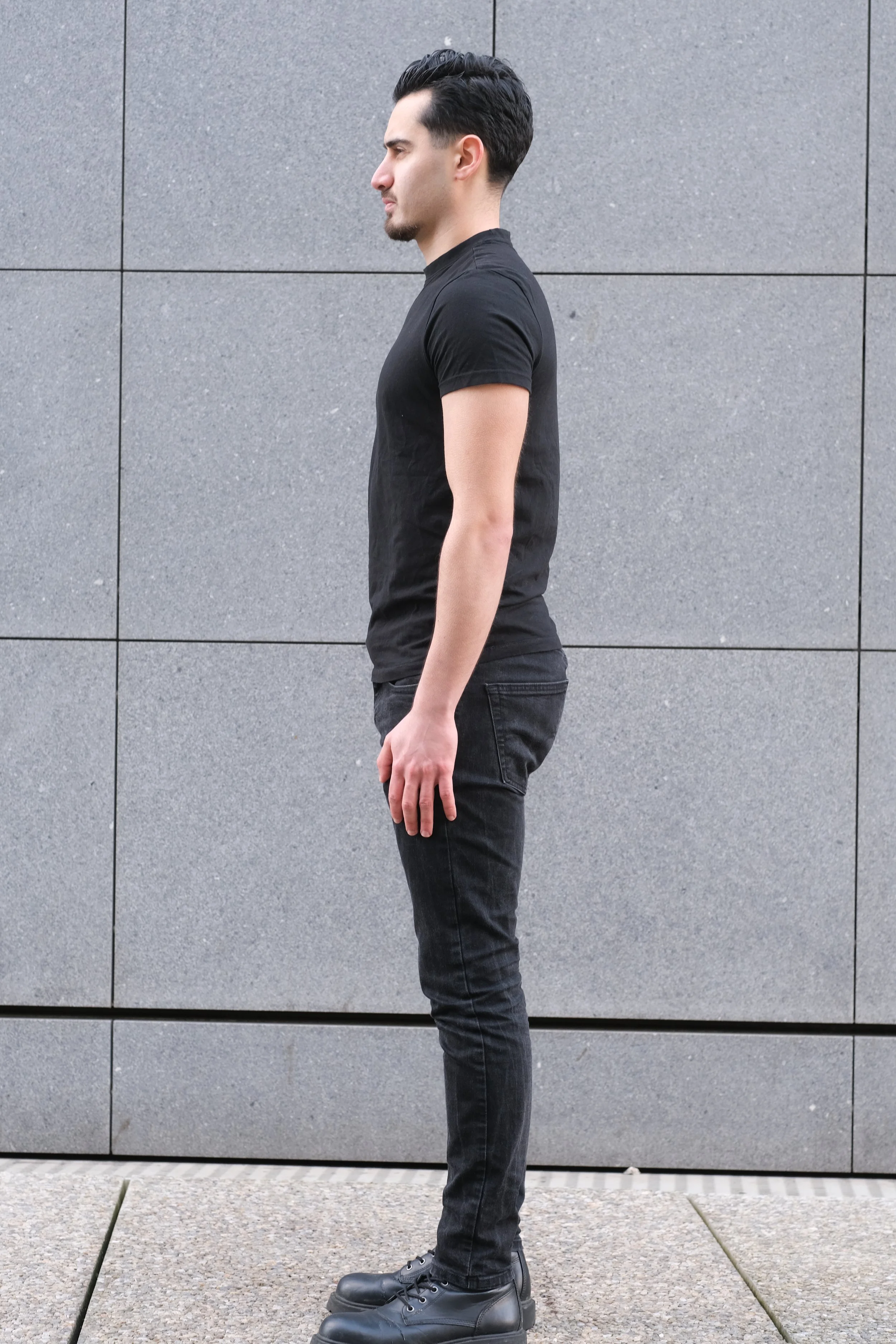 Un homme debout, vu de profil, porte un t-shirt noir, des pantalons noirs et des bottes noires, se tenant devant un mur gris en pierre.