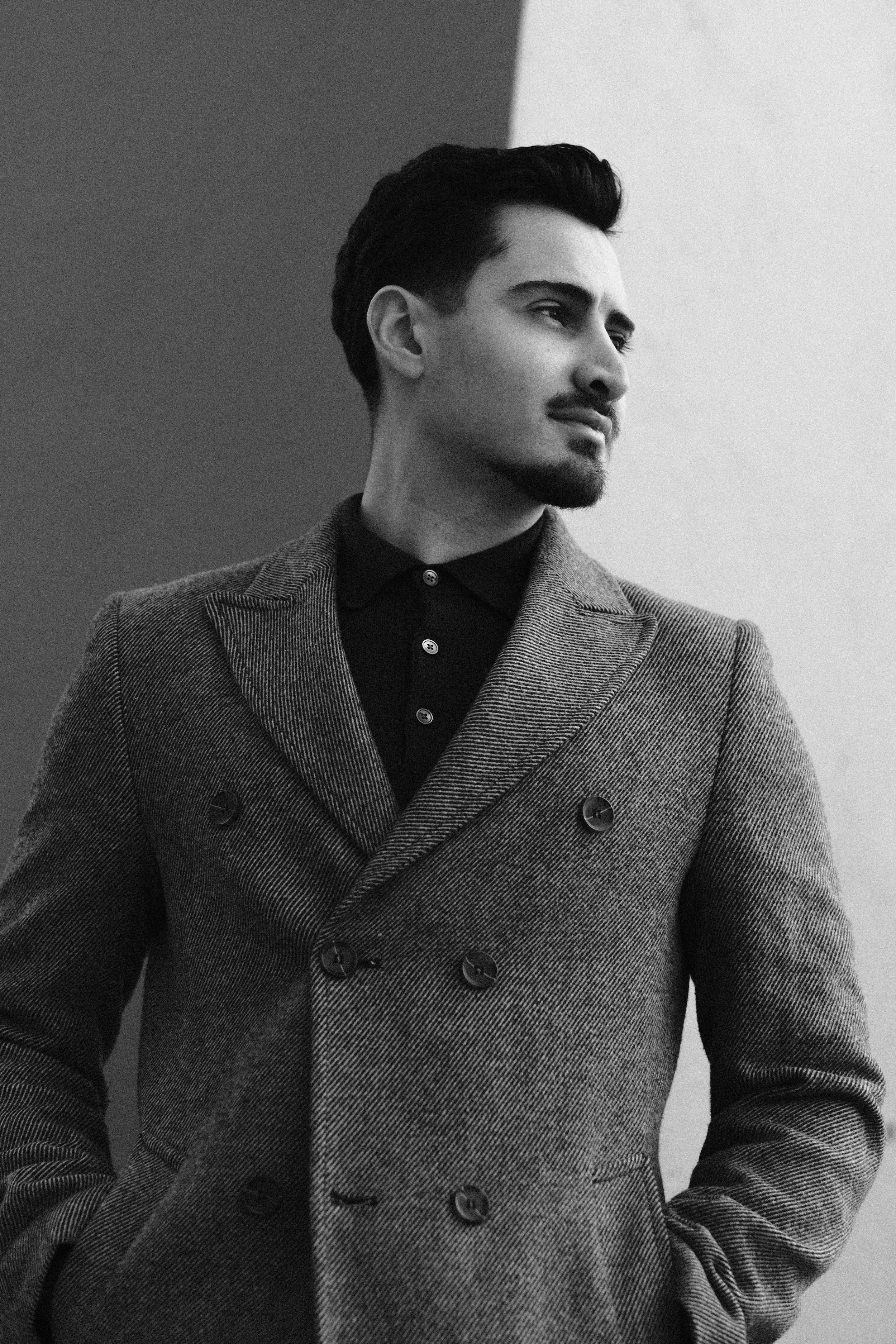Un jeune homme élégant, avec des cheveux noirs et une barbe, portant un manteau en tweed, regarde à gauche dans une photo en noir et blanc.
