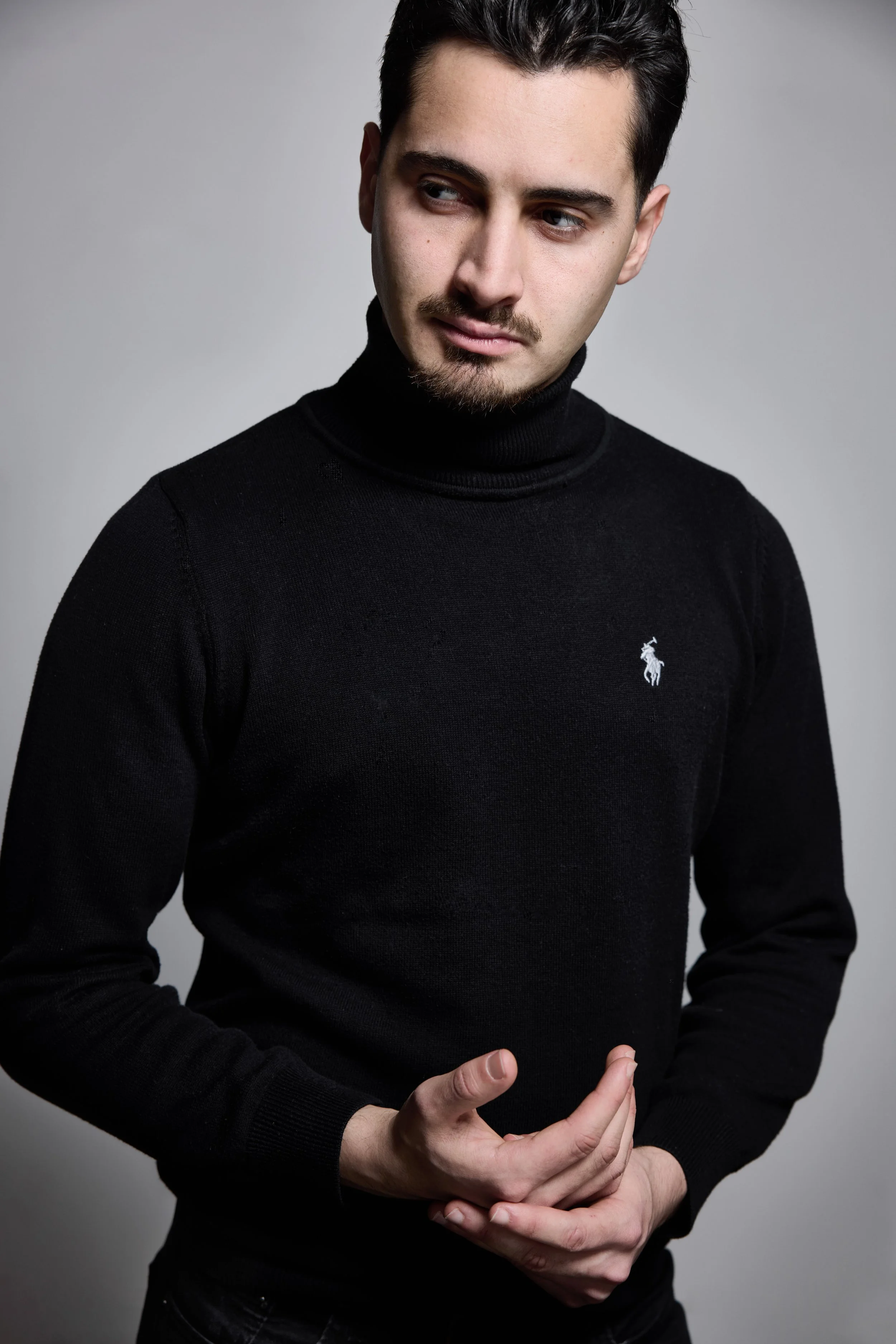 Jeune homme portant un pull noir avec un logo de cheval blanc, poing fermé, regardant sur le côté, fond gris.