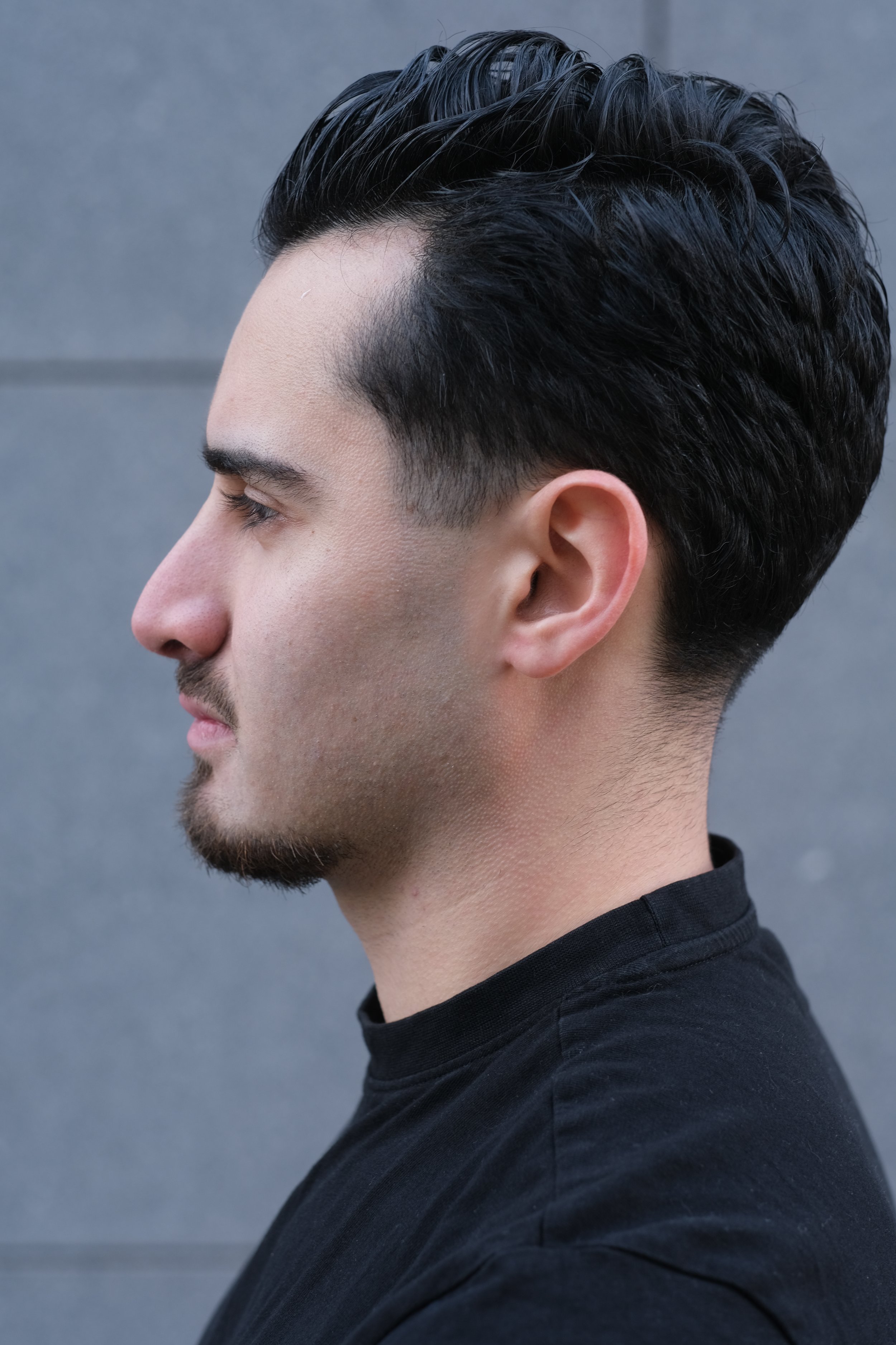 Profil d'un homme avec une coupe de cheveux courte sur les côtés et plus longue sur le dessus, portant un t-shirt noir, contre un fond gris.