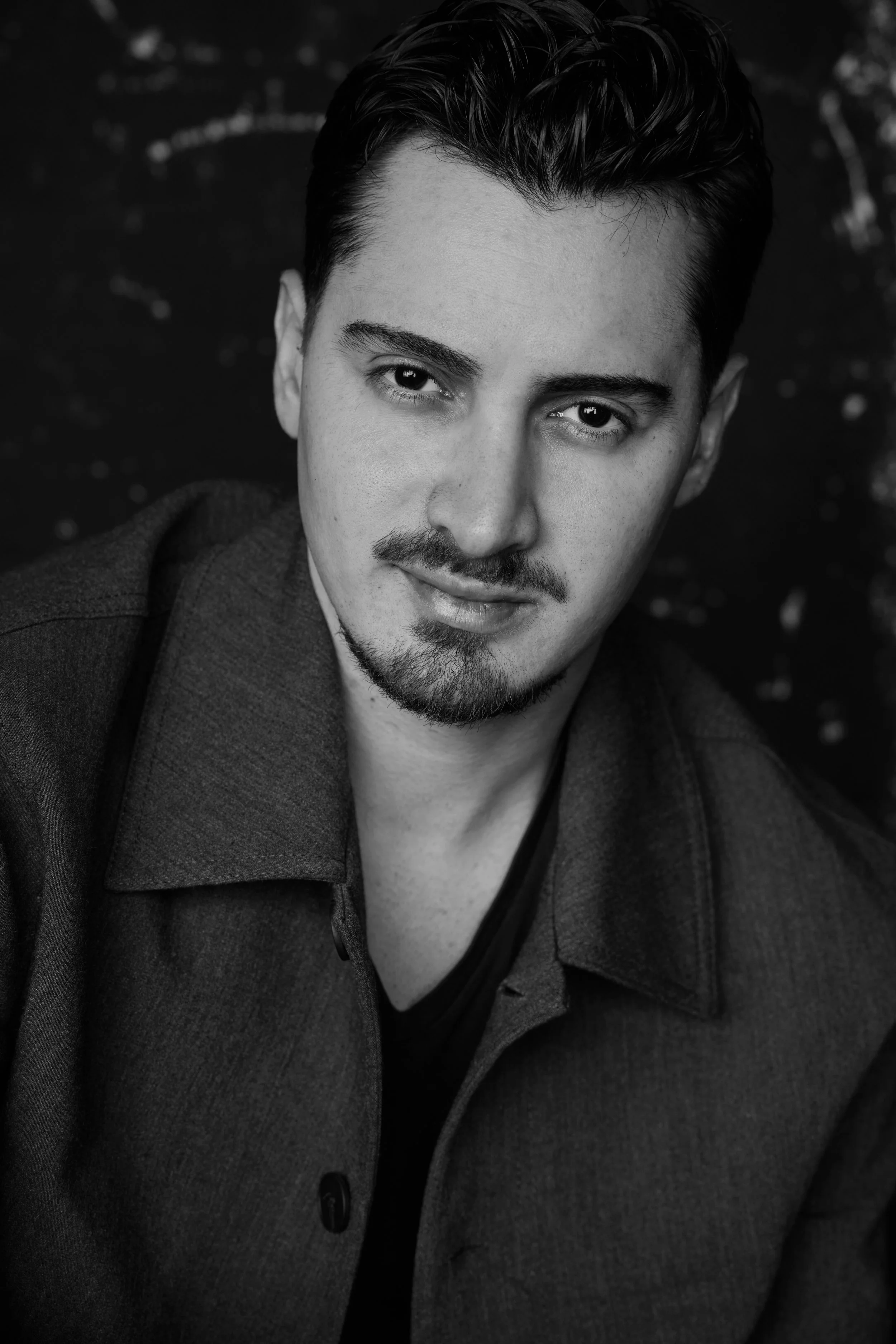 Portrait en noir et blanc d'un jeune homme avec des cheveux foncés, portant un manteau et une chemise. Son regard est dirigé vers l'objectif, avec un léger sourire.