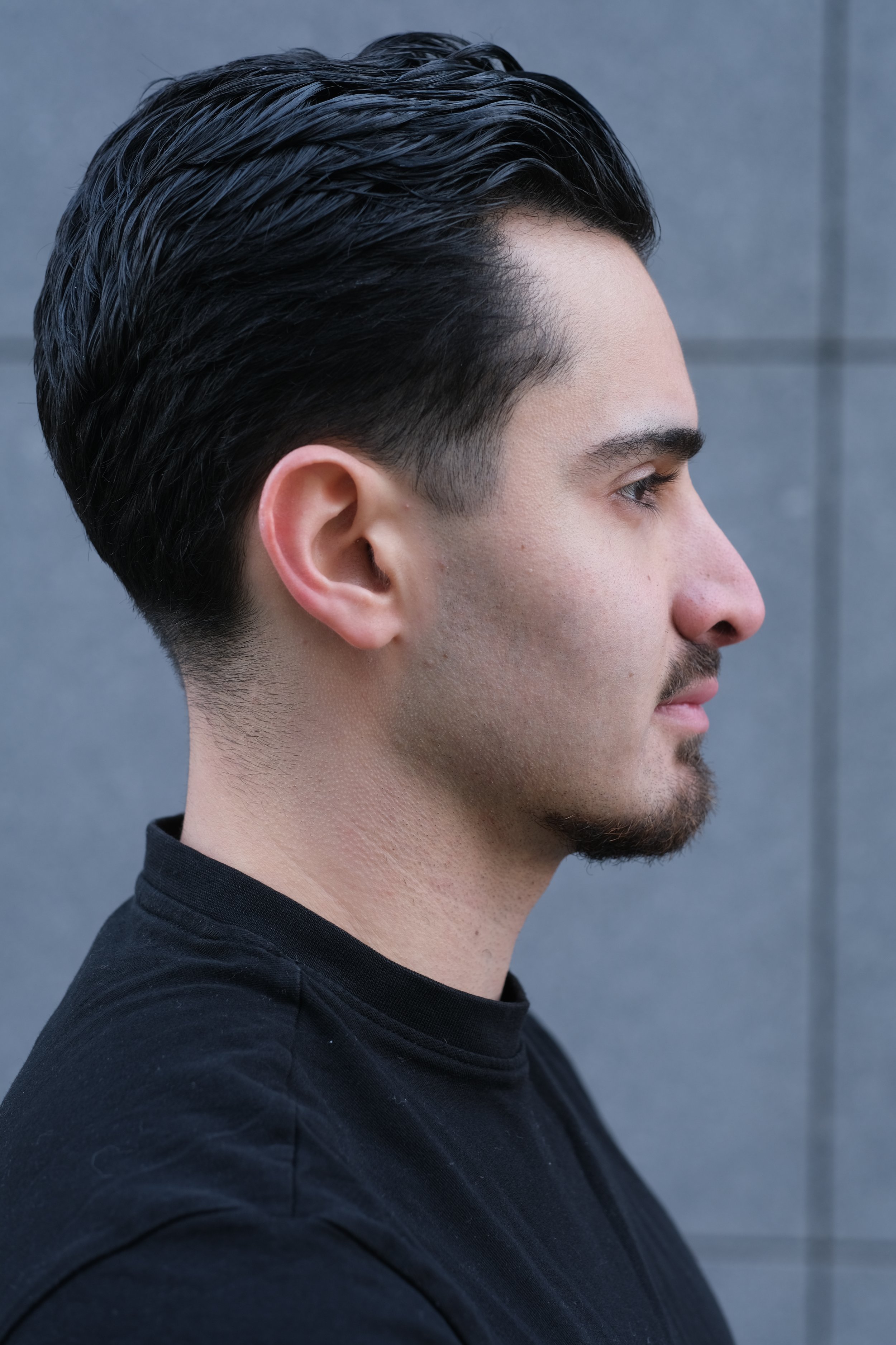 Profil d'un homme avec des cheveux noirs, courts sur les côtés et longs sur le dessus, portant un t-shirt noir, contre un fond gris.
