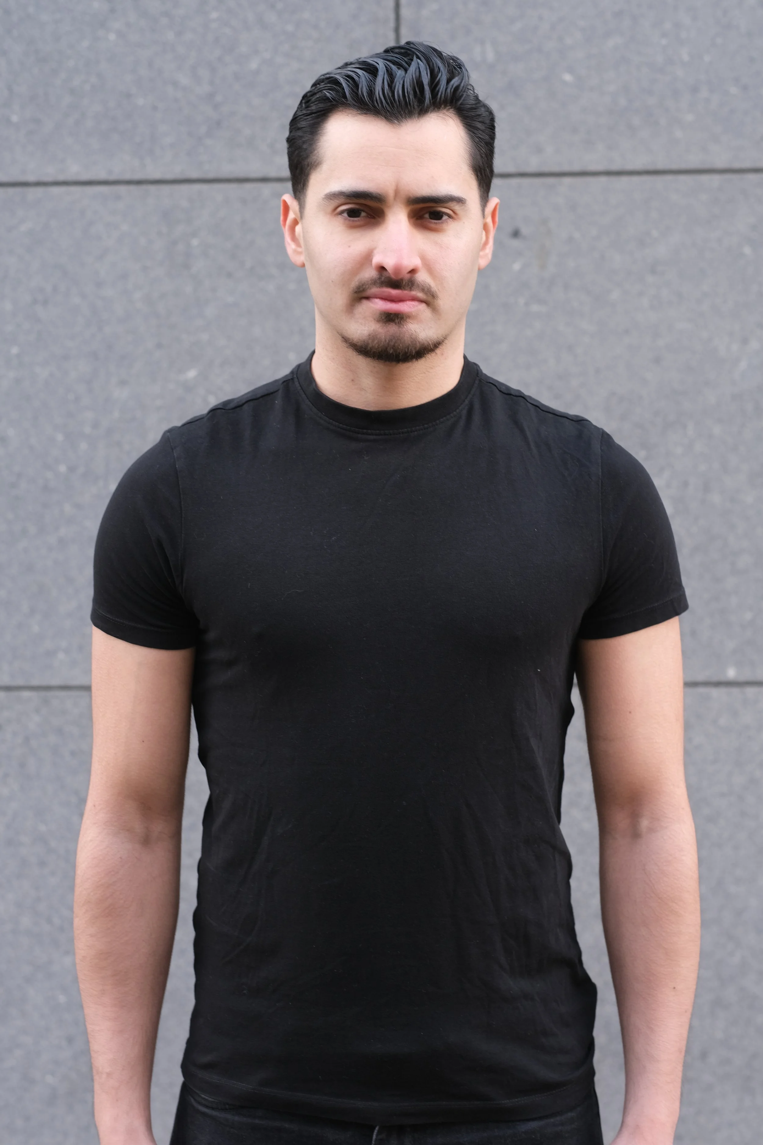 Un homme avec des cheveux noirs, une moustache et une barbe légère, portant un t-shirt noir, se tient devant un mur gris.