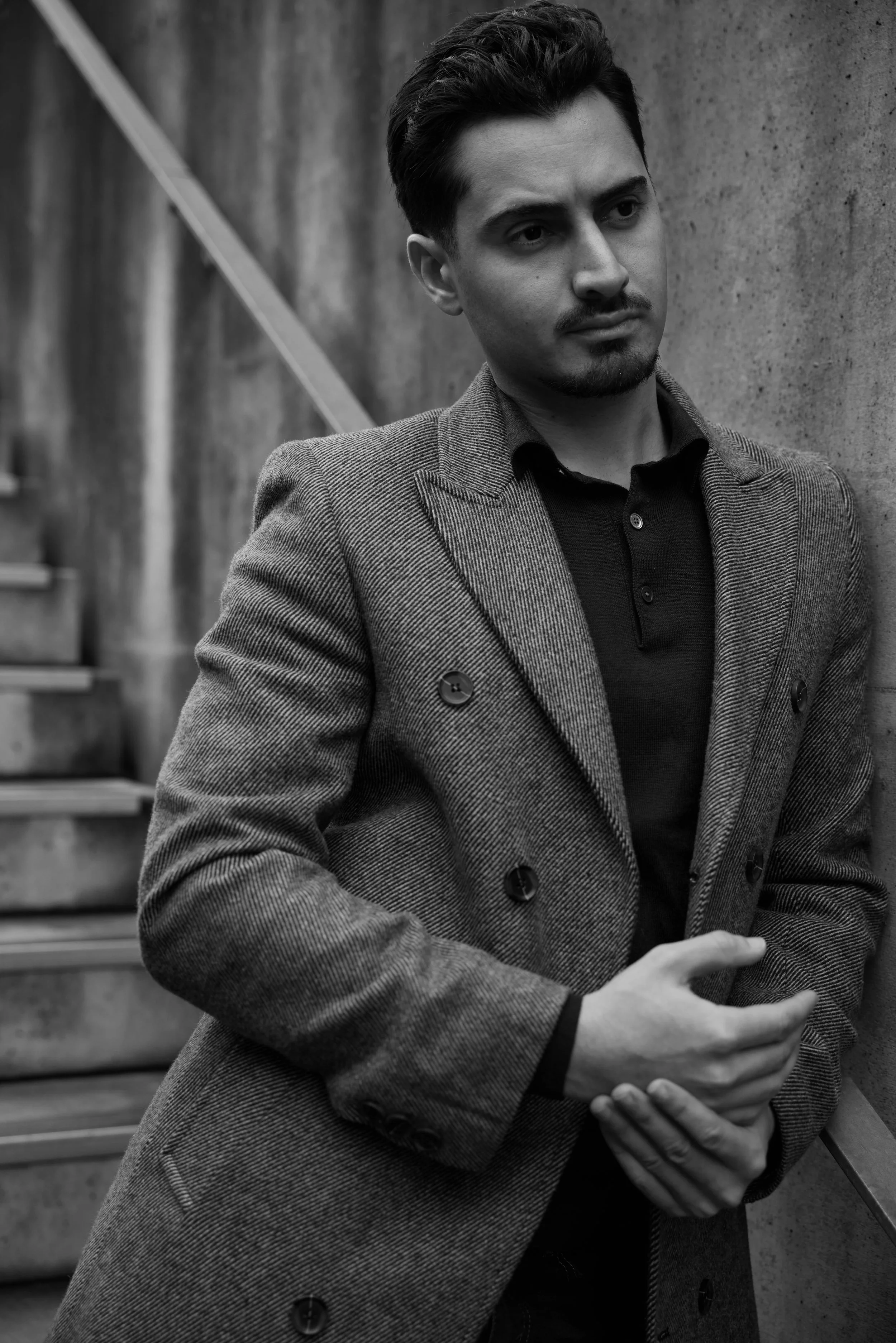 Un homme portant un blazer à carreaux et une chemise noire, avec une moustache, est dans un cadre urbain minimaliste. Il regarde pensivement vers la droite, ses bras croisés devant lui.