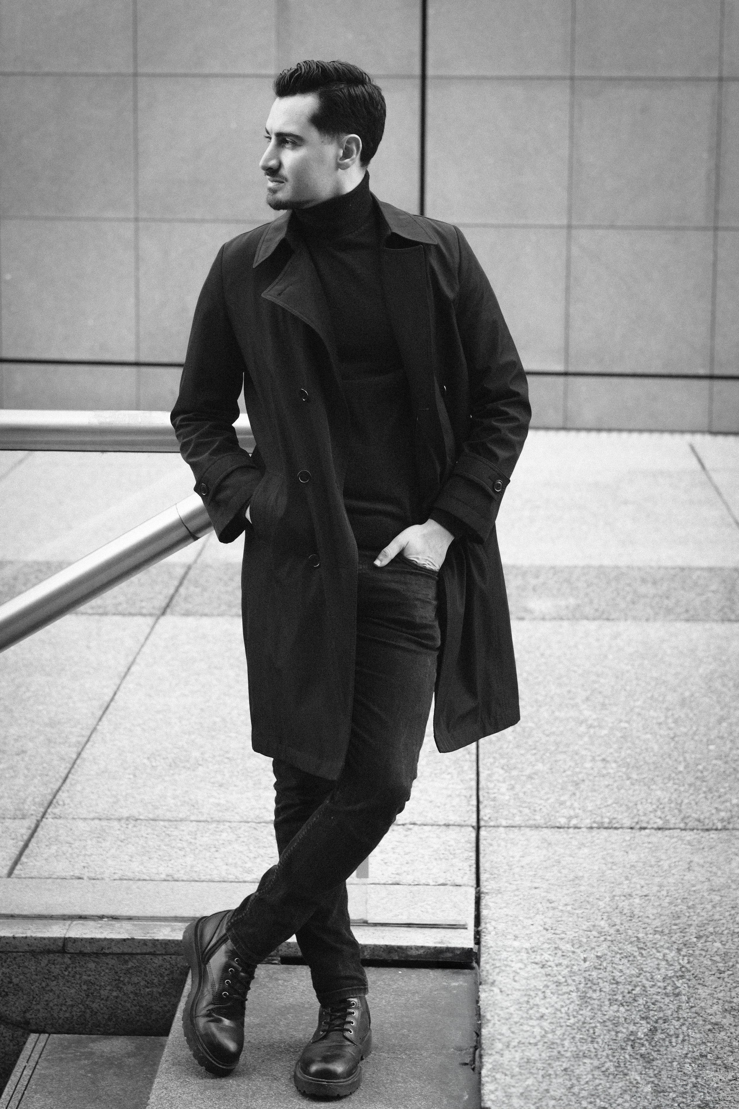 Un homme en vêtements noirs, portant un trench coat et des bottes, se tient dans un environnement urbain devant un mur en carreaux, avec une main dans la poche.
