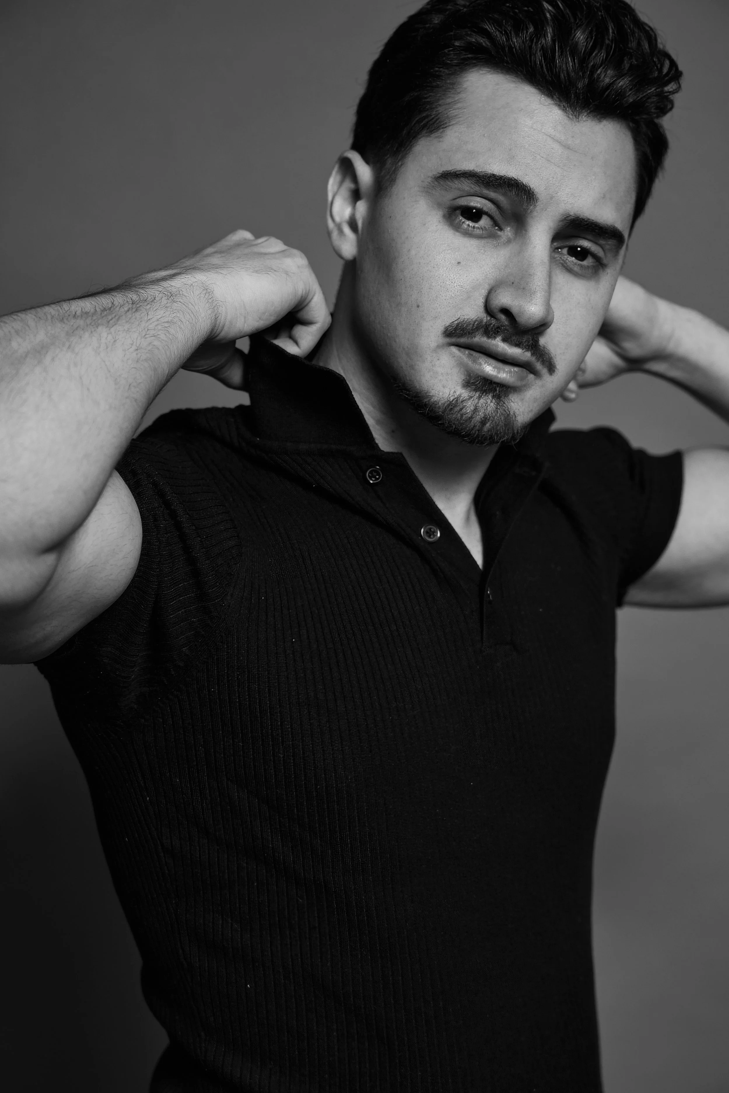 Portrait en noir et blanc d'un jeune homme avec des cheveux foncés, une barbe légère et un regard pensif, portant un polo noir avec des bras levés derrière la tête.