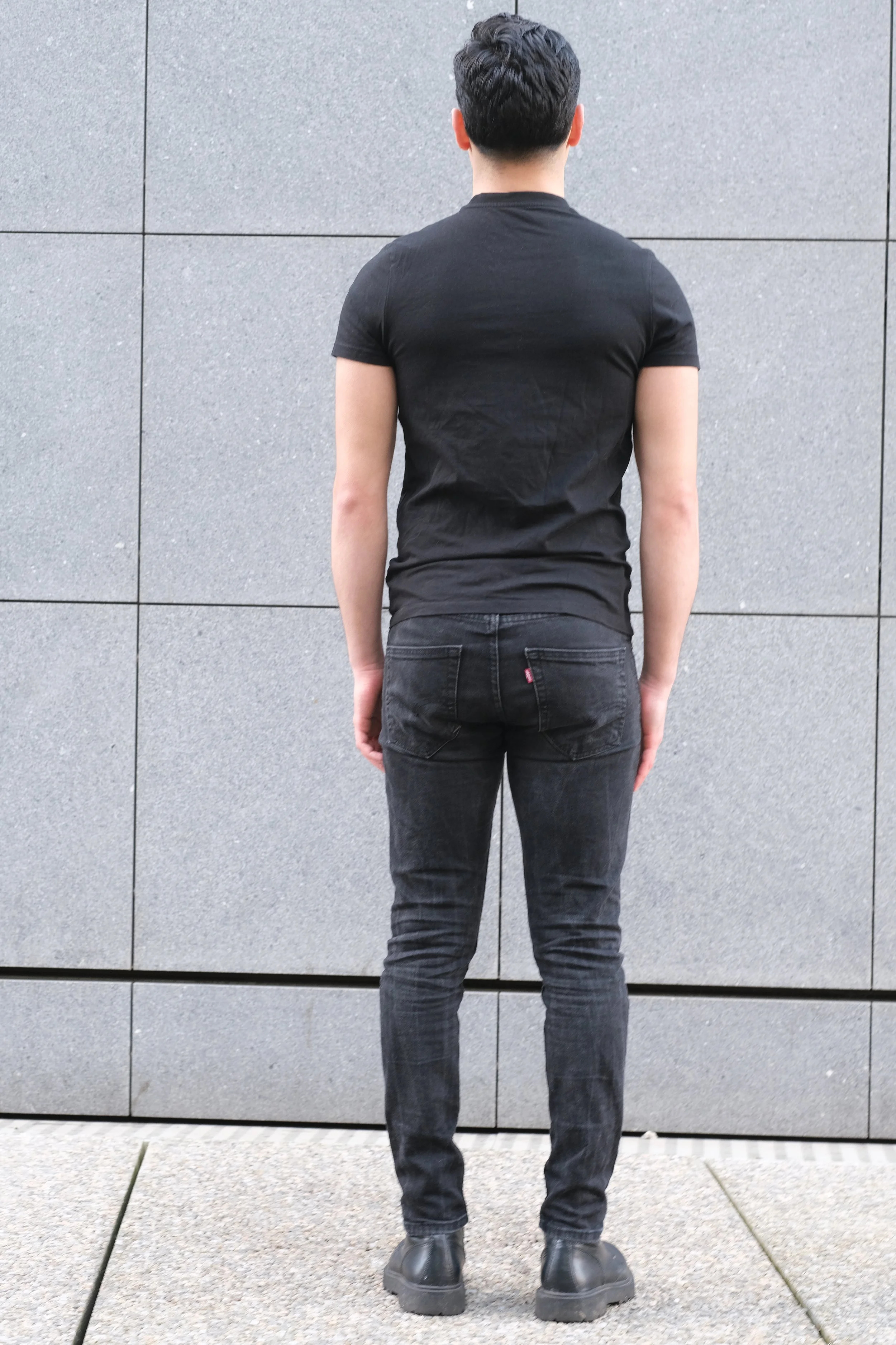 Une personne debout de dos portant un t-shirt noir, un pantalon noir et des chaussures noires, se tenant contre un mur de pierre gris.