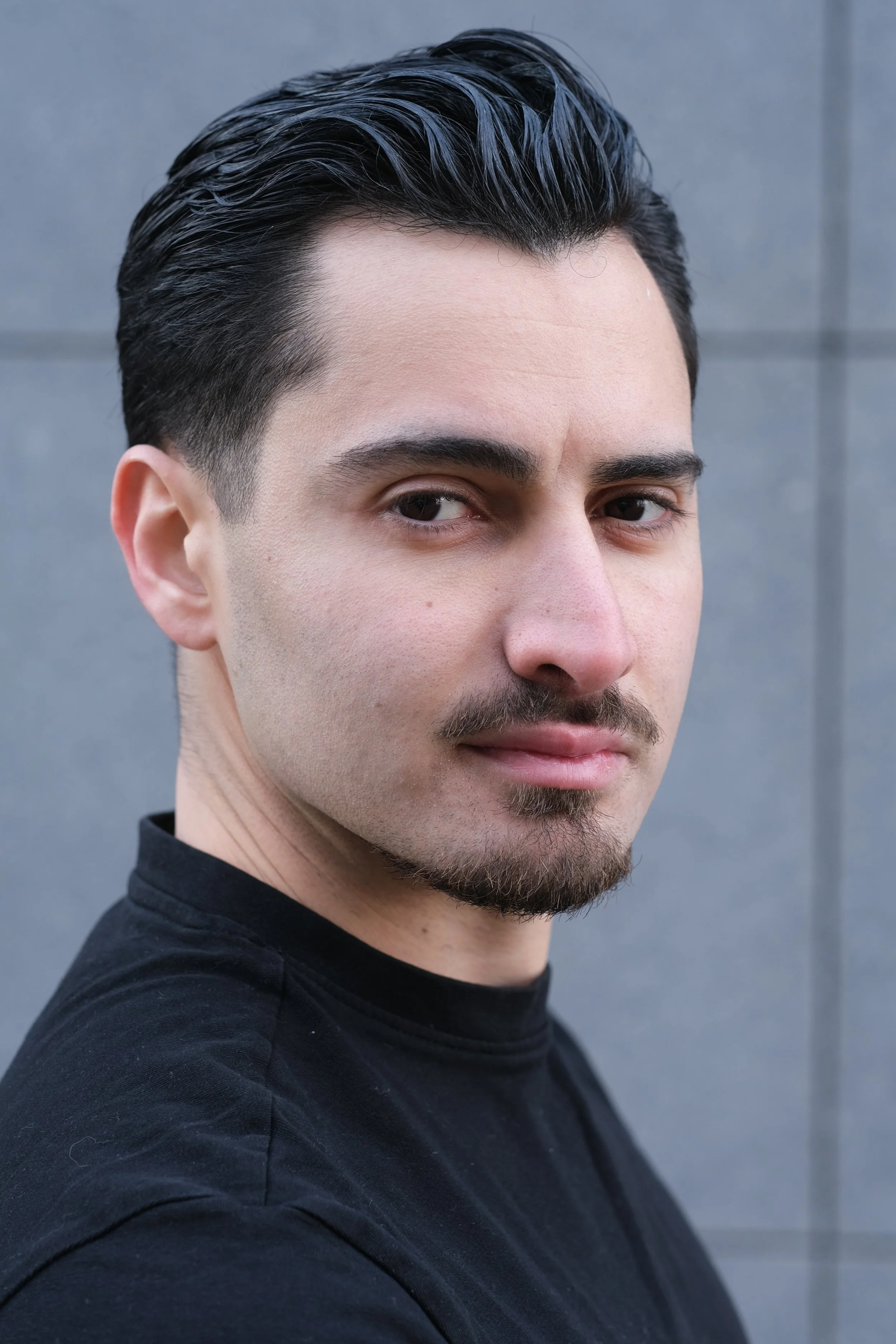 Portrait d'un homme avec une coiffure noire, barbe légère, portant un t-shirt noir, devant un mur gris.