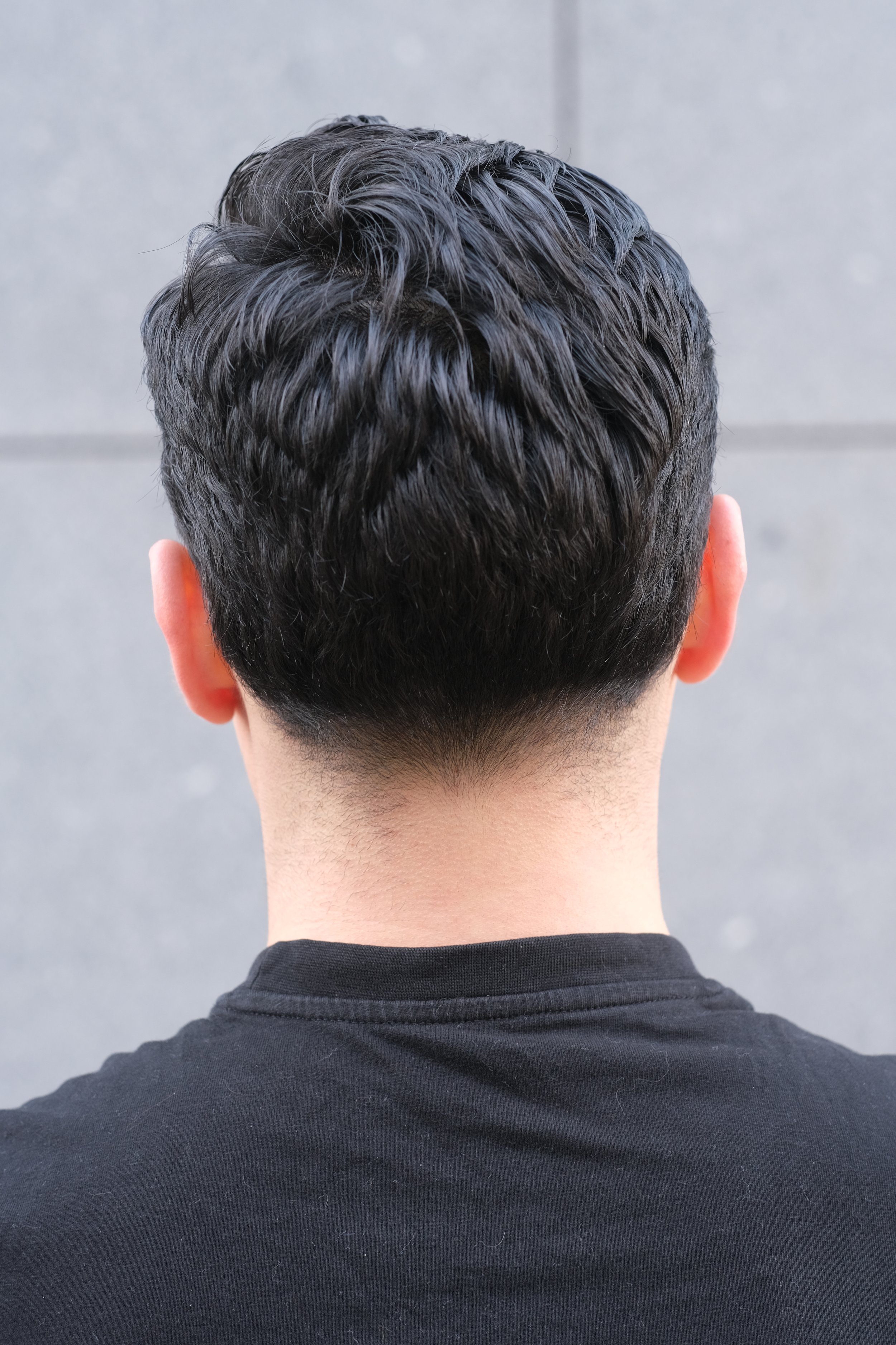 Vue de derrière d'une personne avec cheveux courts et bouclés, portant un t-shirt noir, contre un mur gris.