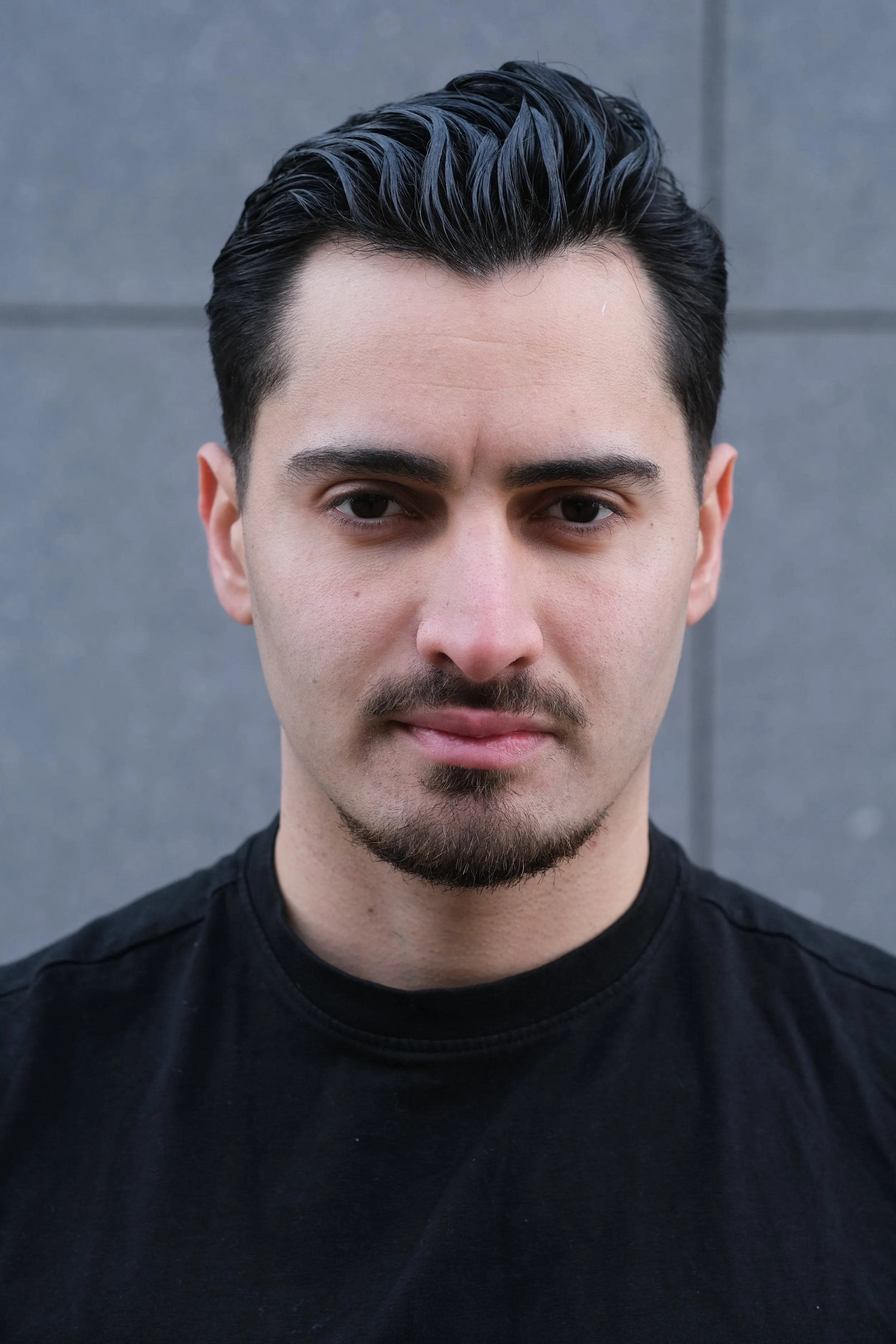 Jeune homme avec cheveux noirs coiffés en arrière, portant un t-shirt noir, se tenant devant un mur gris.