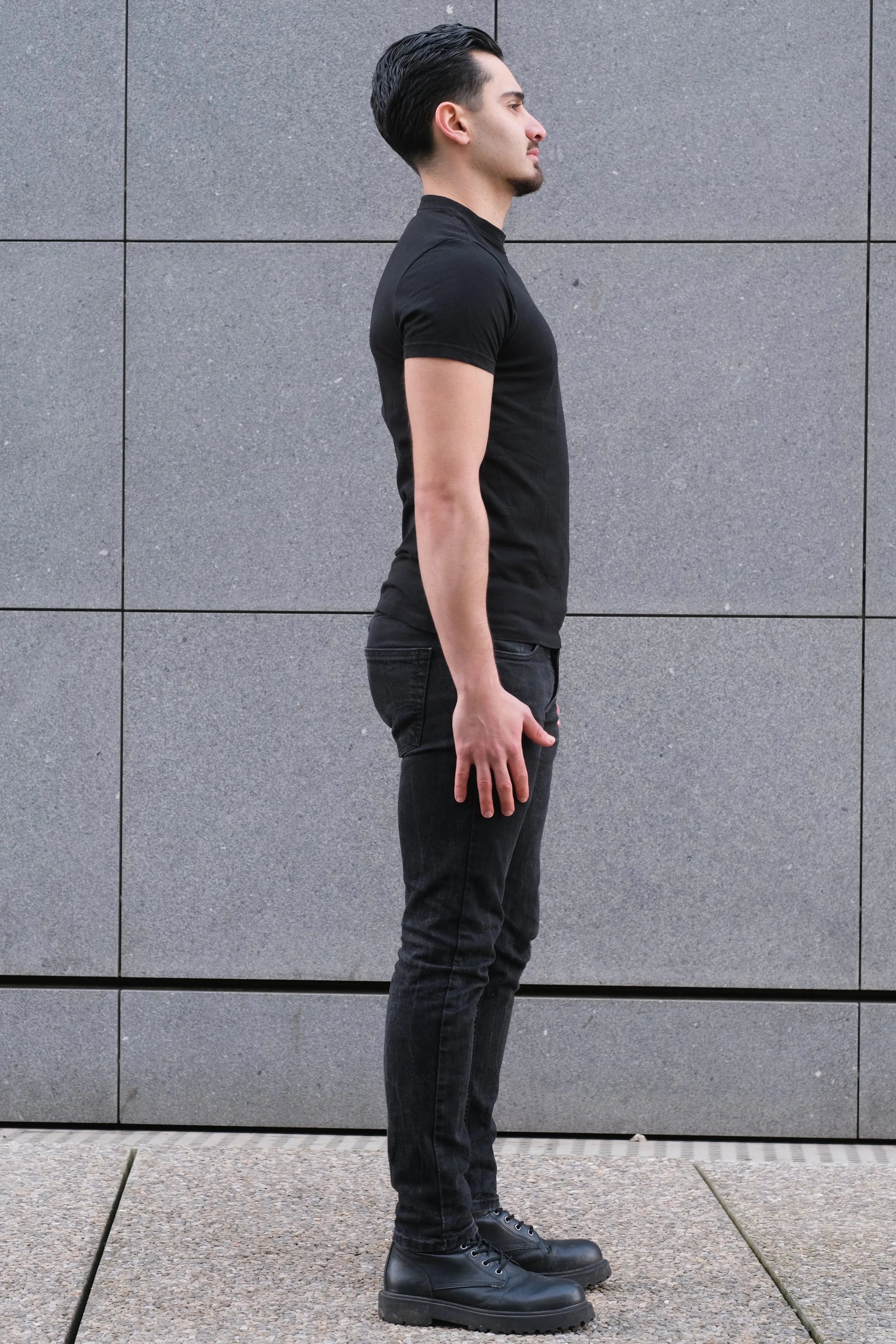 Jeune homme de profil portant une tenue noire, debout contre un mur en pierre grise.