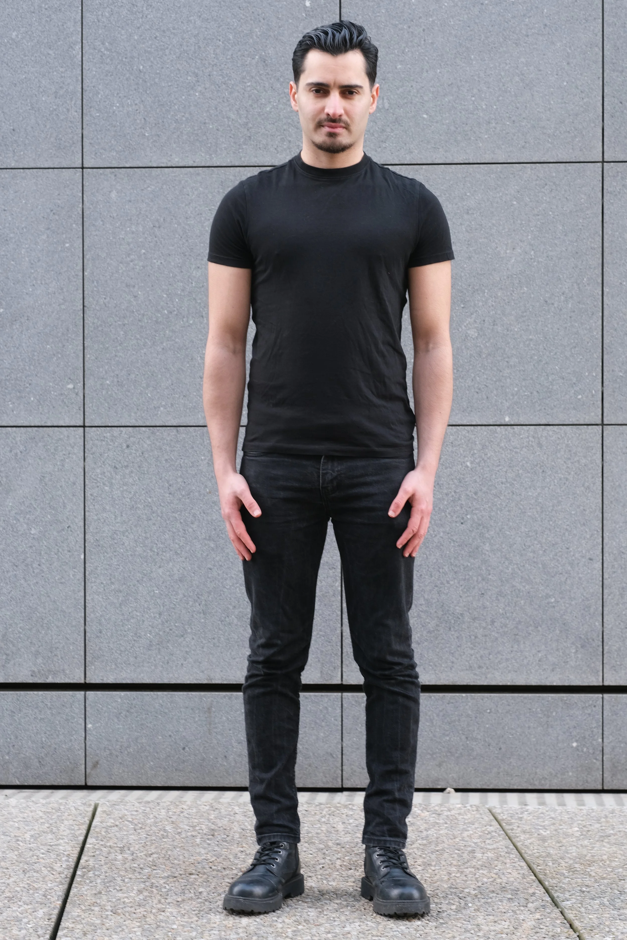 Un jeune homme avec des cheveux noirs, portant un t-shirt noir, un pantalon noir, et des bottes noires, se tient devant un mur de pierre grise. Il regarde l'objectif avec une expression neutre.