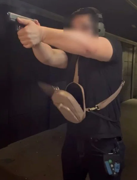 Person aiming a handgun with both hands in a shooting range, wearing a black shirt and a tan crossbody bag.