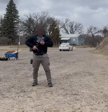 A person standing outside on a gravel lot, holding a gun, with a wheelbarrow and a white car in the background, and trees and a building under cloudy skies.