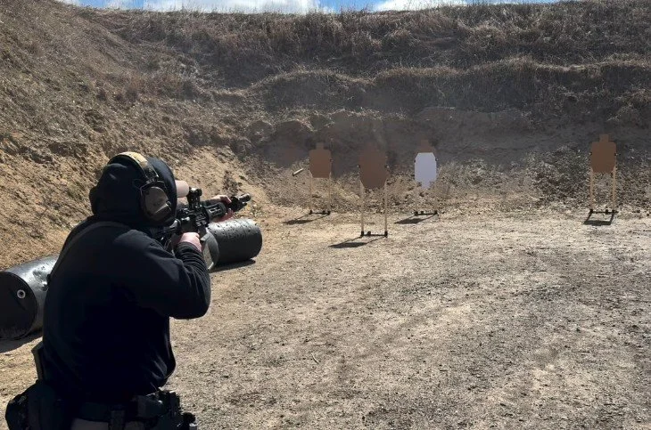 Person at a shooting range aiming a rifle at target silhouettes against a dirt wall.
