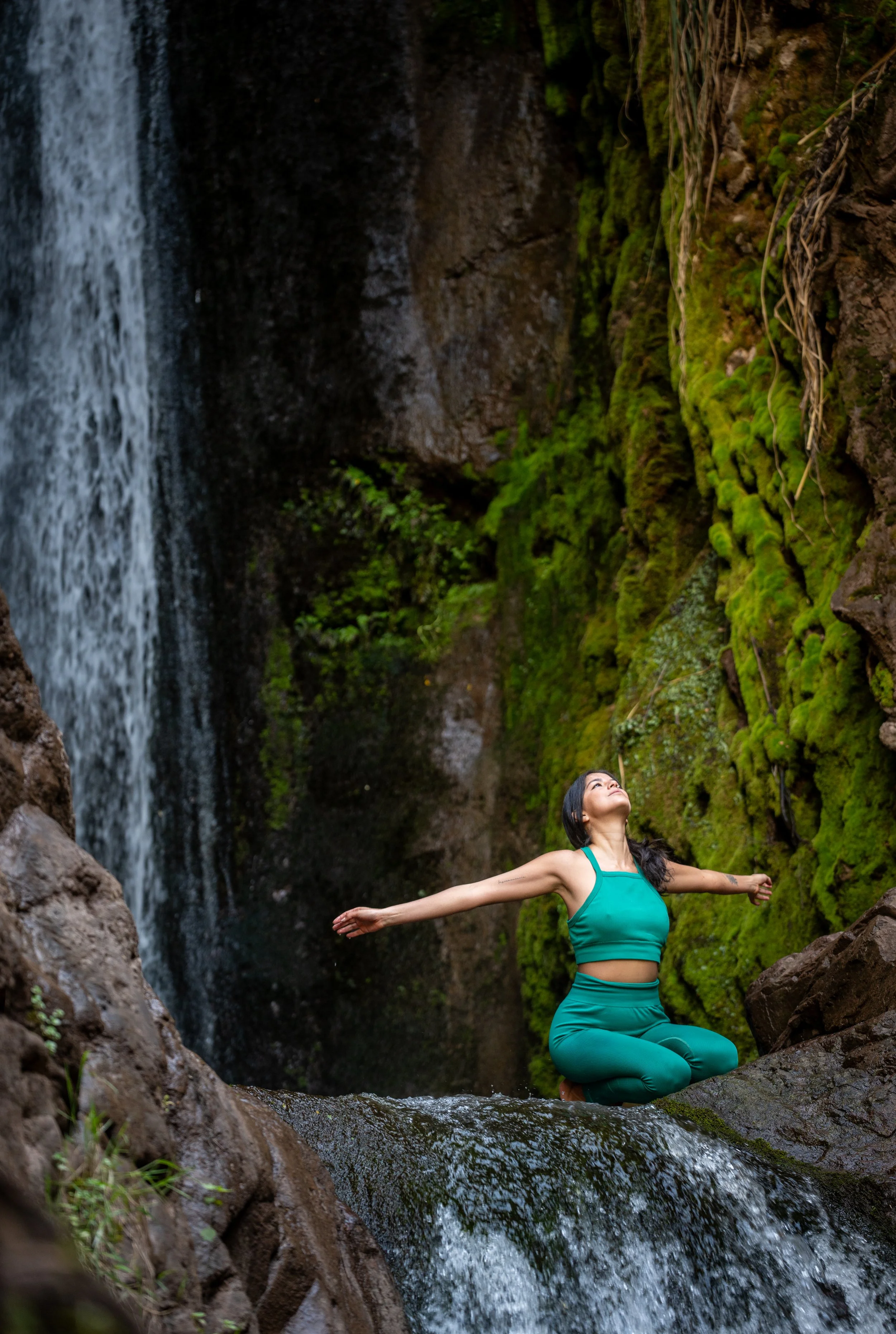 A woman practicing yoga in a tree pose with arms outstretched, kneeling on rocks in front of a waterfall surrounded by moss-covered rocks and greenery.