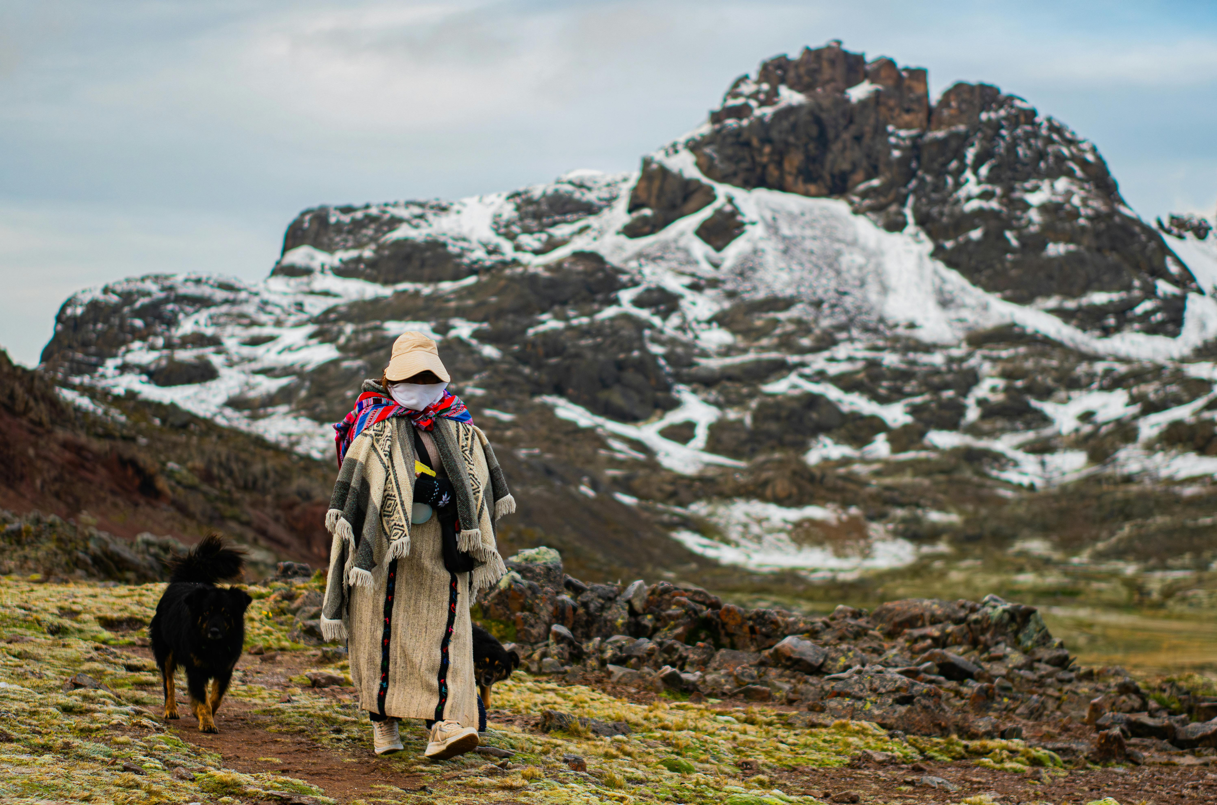 Person wearing a hat, white face covering, and a long patterned coat, walking with two dogs on a trail in a mountainous landscape with snow patches and rugged rock formations.