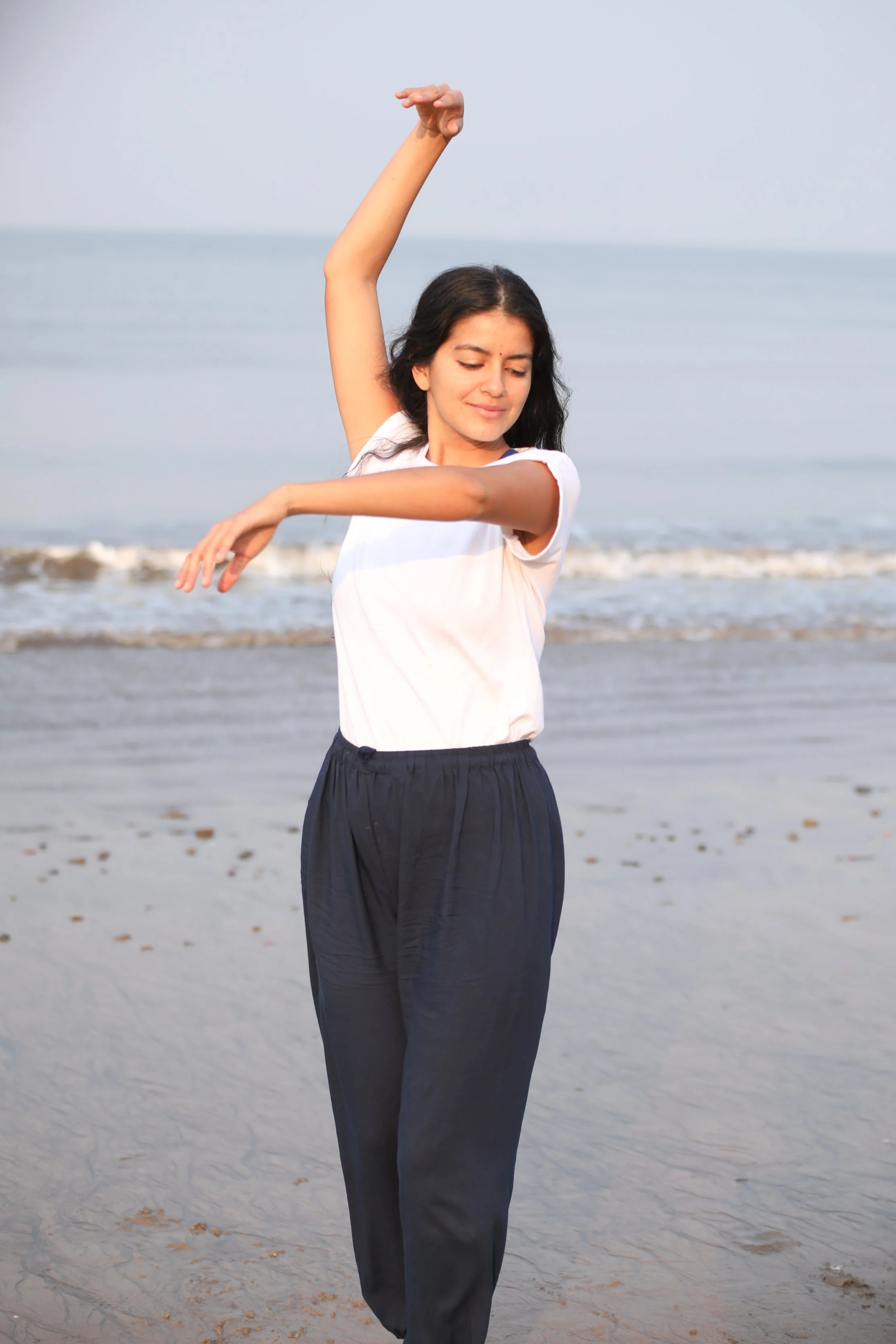 A young woman with dark hair is on a beach, standing with eyes closed, arms raised. One arm is bent over her head, and the other is extended to the side. The ocean waves are in the background.