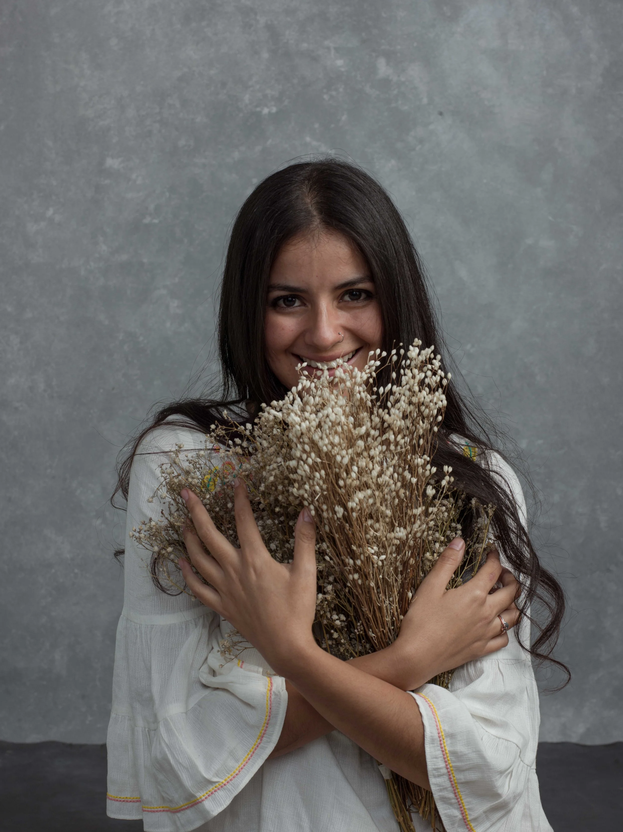 A woman with long dark hair holding a large bouquet of dried flowers in front of her face and chest, smiling. She is wearing a white shirt with colorful embroidery near the sleeves. The background is a gray textured surface.