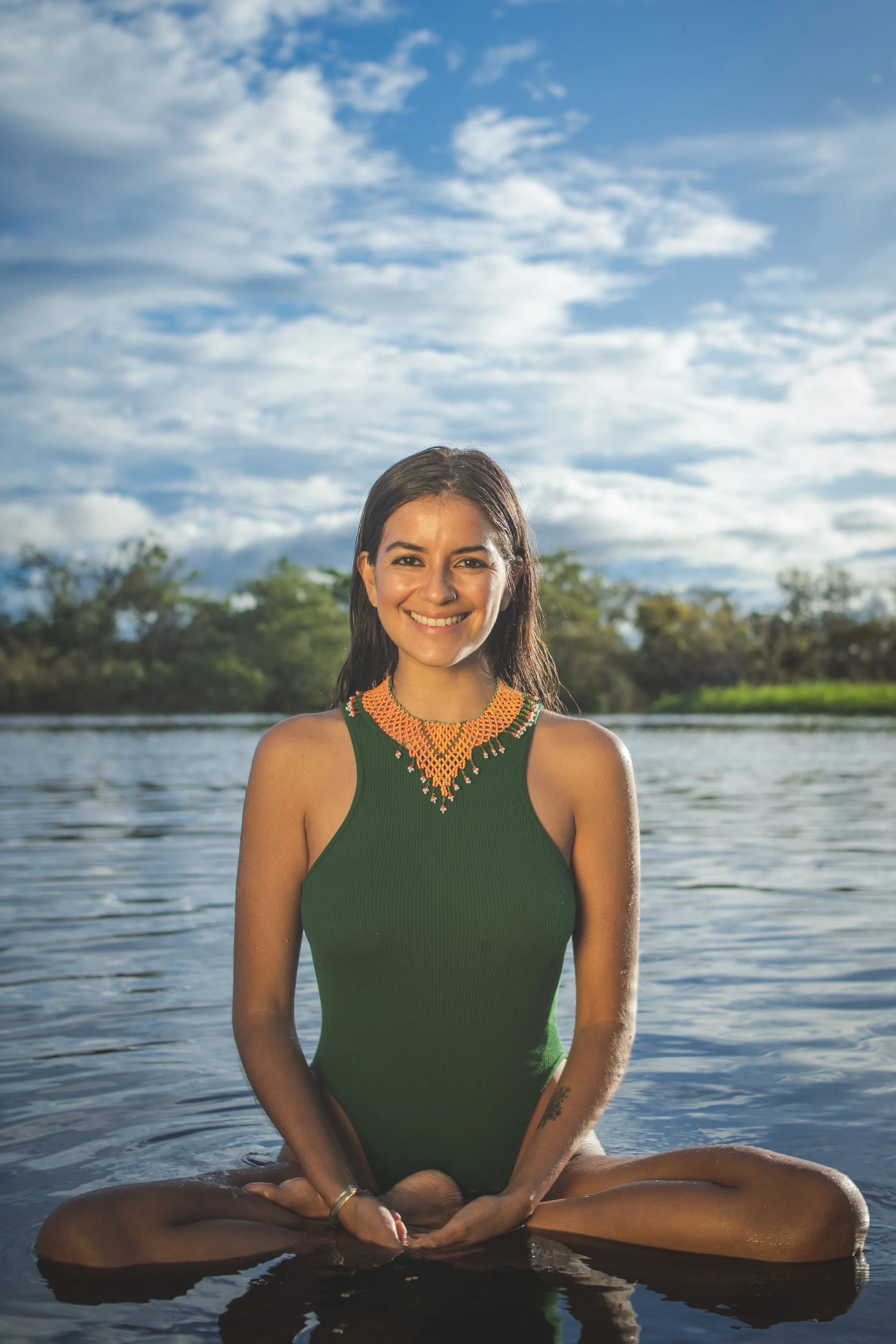 A woman with long dark hair sits cross-legged in shallow water during the daytime, smiling at the camera, with a backdrop of trees and a partly cloudy sky.