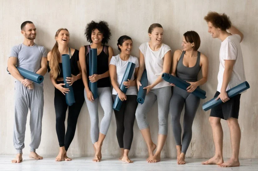 Group of seven diverse people holding yoga mats and smiling together, standing barefoot against a plain wall.