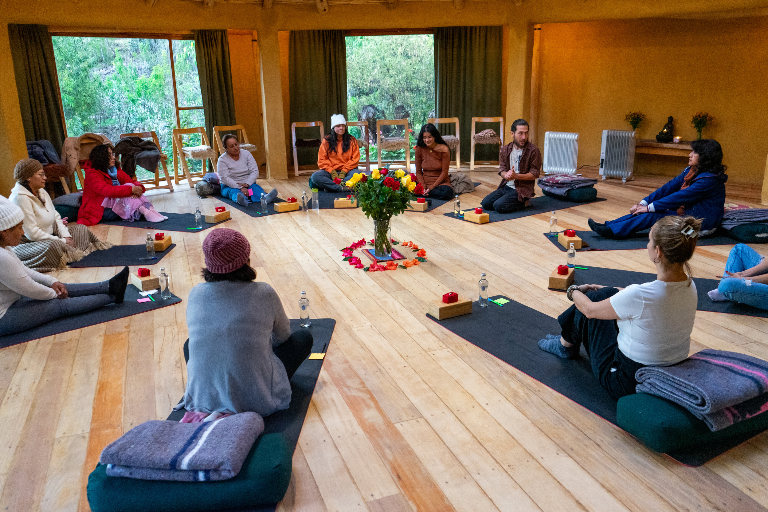 A group of people sitting on mats in a circle in a room with large windows showing greenery outside. A table with a vase of flowers is in the center. The room has wooden floors and walls, with some folded blankets and personal items around.